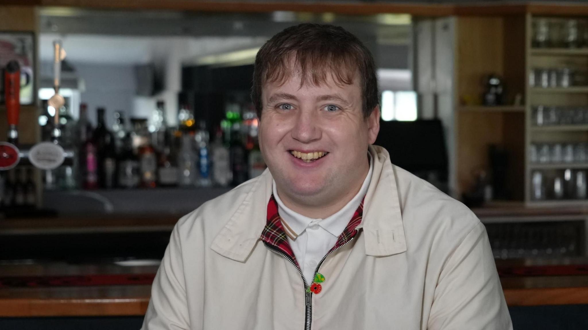Macaulay Robinson sitting in the clubhouse at Kirkley and Pakefield Football Club in Lowestoft in front of the old wooden style bar. He is wearing a beige scooter style jacket with a poppy on it and a tartan lining. He is wearing a white polo shirt and has a brown fringe. He has a wide smile and is of stocky build.