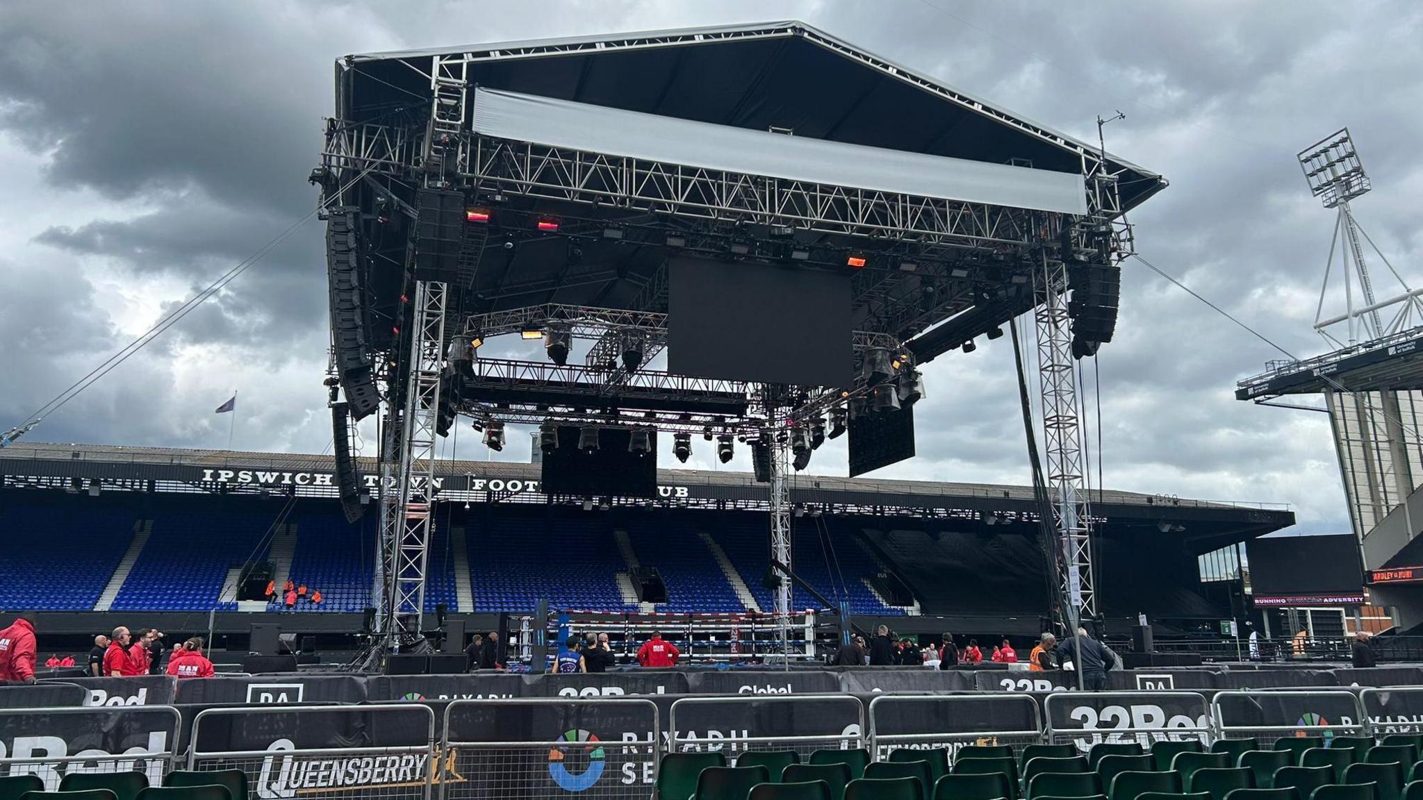 A view of the boxing ring in the centre of the Portman Road pitch from a behind the first couple of rows of green seats. Behind the boxing ring is one of the stands at the stadium.