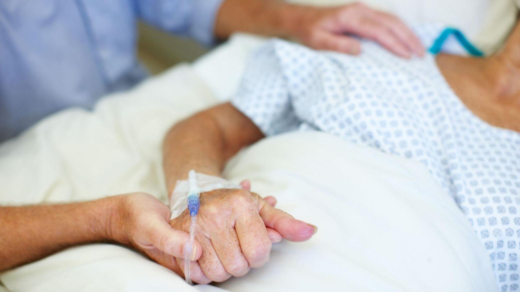 An older woman wearing a hospital gown, lying in a hospital bed with a IV inserted into her hand. A man in a blue shirt sits beside her, holding her hand in his, while his other hand rests on her shoulder. 