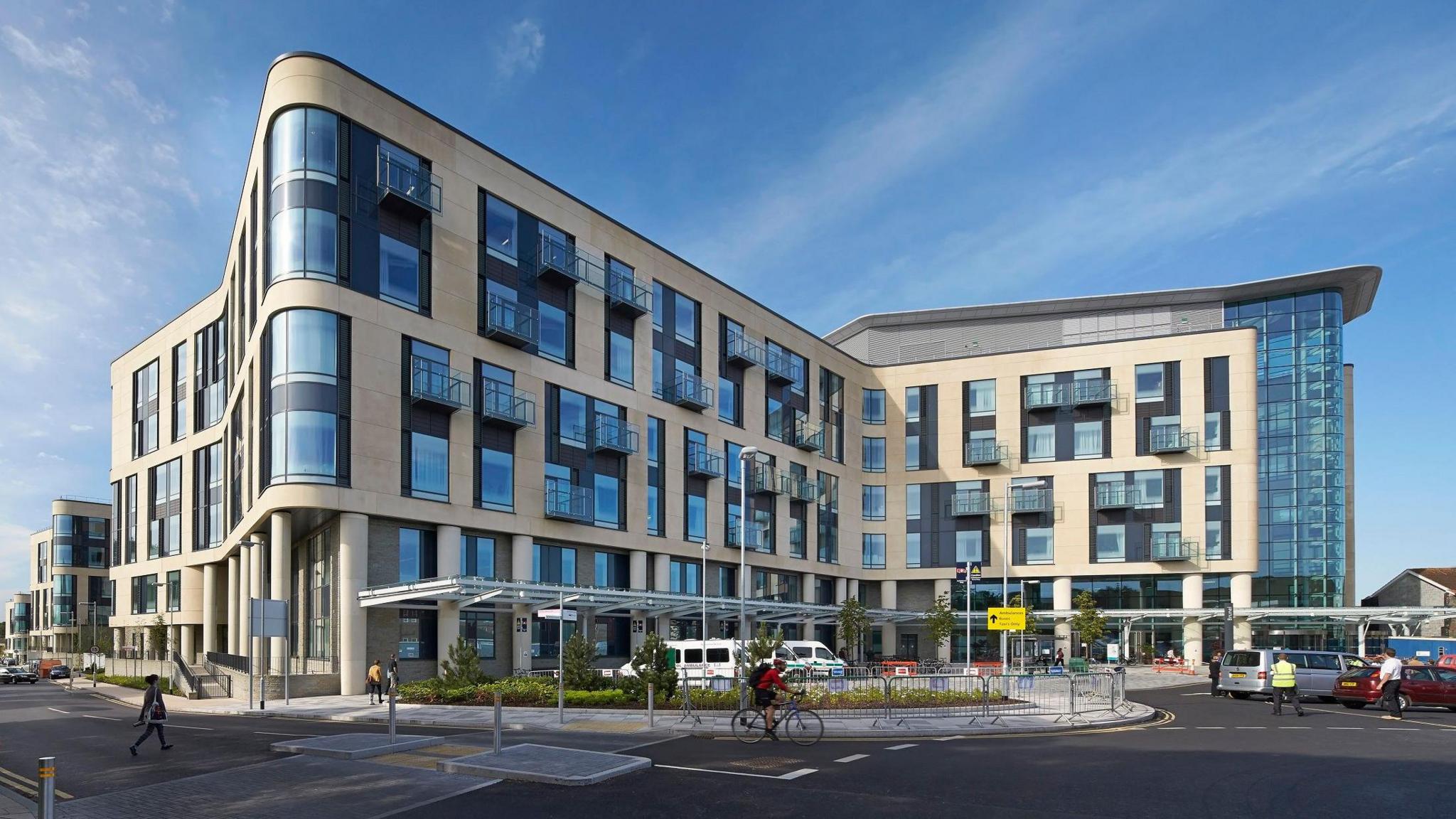 An exterior shot of the main entrance to Southmead Hospital in Bristol. The exterior of the building is of a modern style with the building's edges curved and large windows. There are several vehicles parked outside and pedestrians walking towards the building