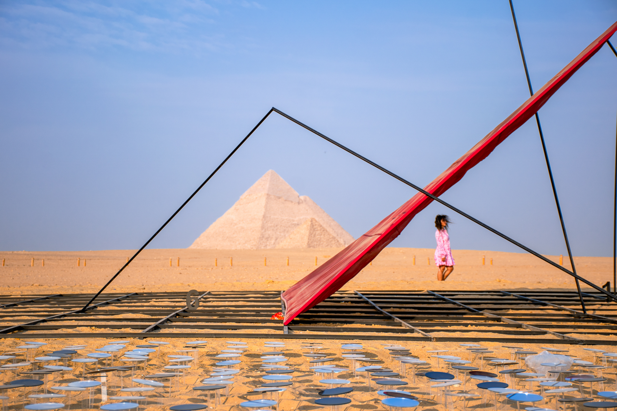 The Great Pyramid stands in the background on the sand. It is framed by a modern metal structure.