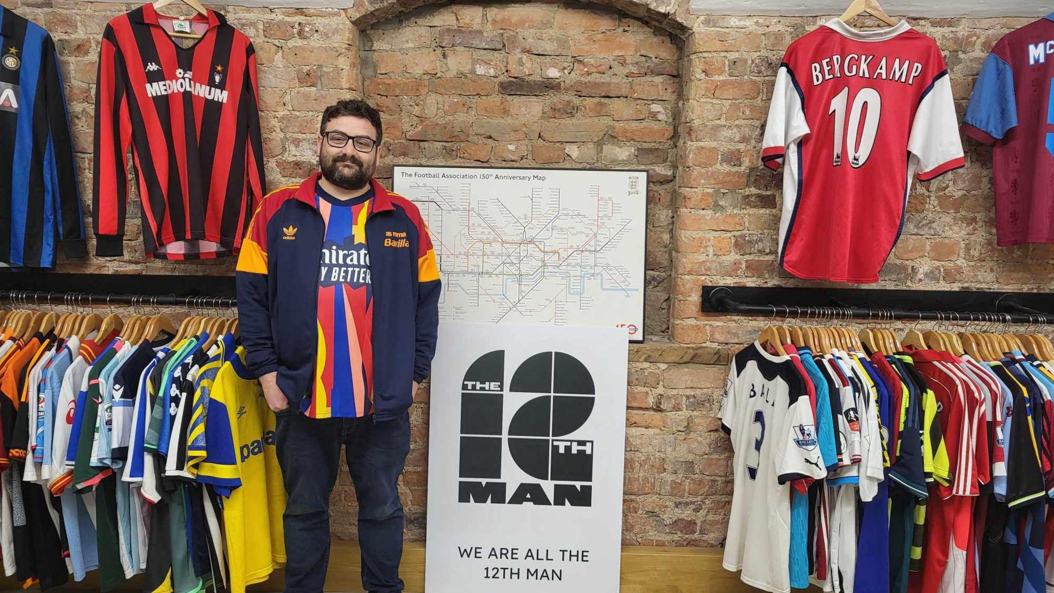 Shaun Owen has black hair and a black beard and moustache. He is wearing glasses and a red, yellow and blue retro football shirt, with a navy, red and yellow jacket on top. He is standing in front of a brick wall next to a sign that says "The 12th man". Either side of him on the wall are hangers holding dozens of retro football shirts. 