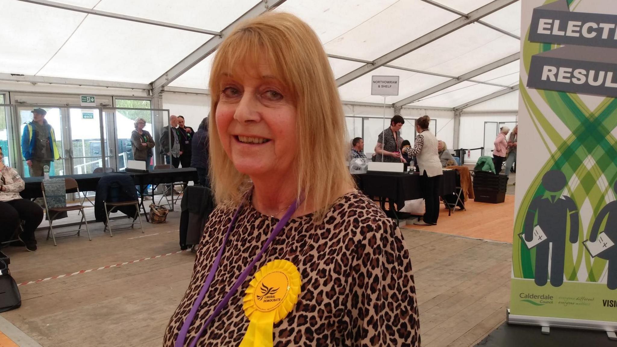 A woman smiling at the camera inside a marquee during a local election vote count. She has straight, shoulder-length, strawberry blonde hair and is wearing a leopard print top adorned with a yellow Liberal Democrats rosette. In the background are several people standing and sitting behind a row of tables.