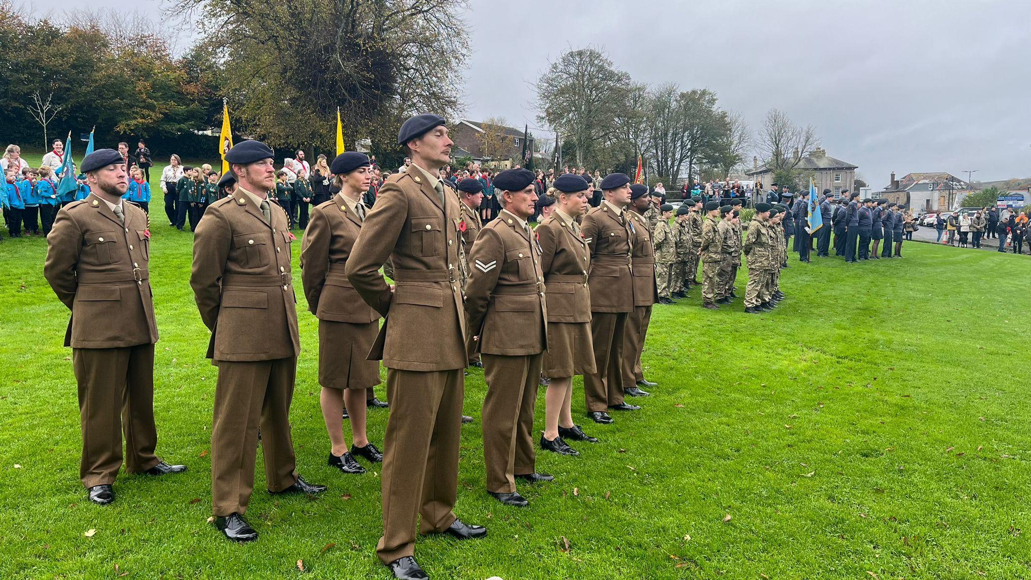 Soldiers in uniform stand in rows, on grass, with children grouped behind them