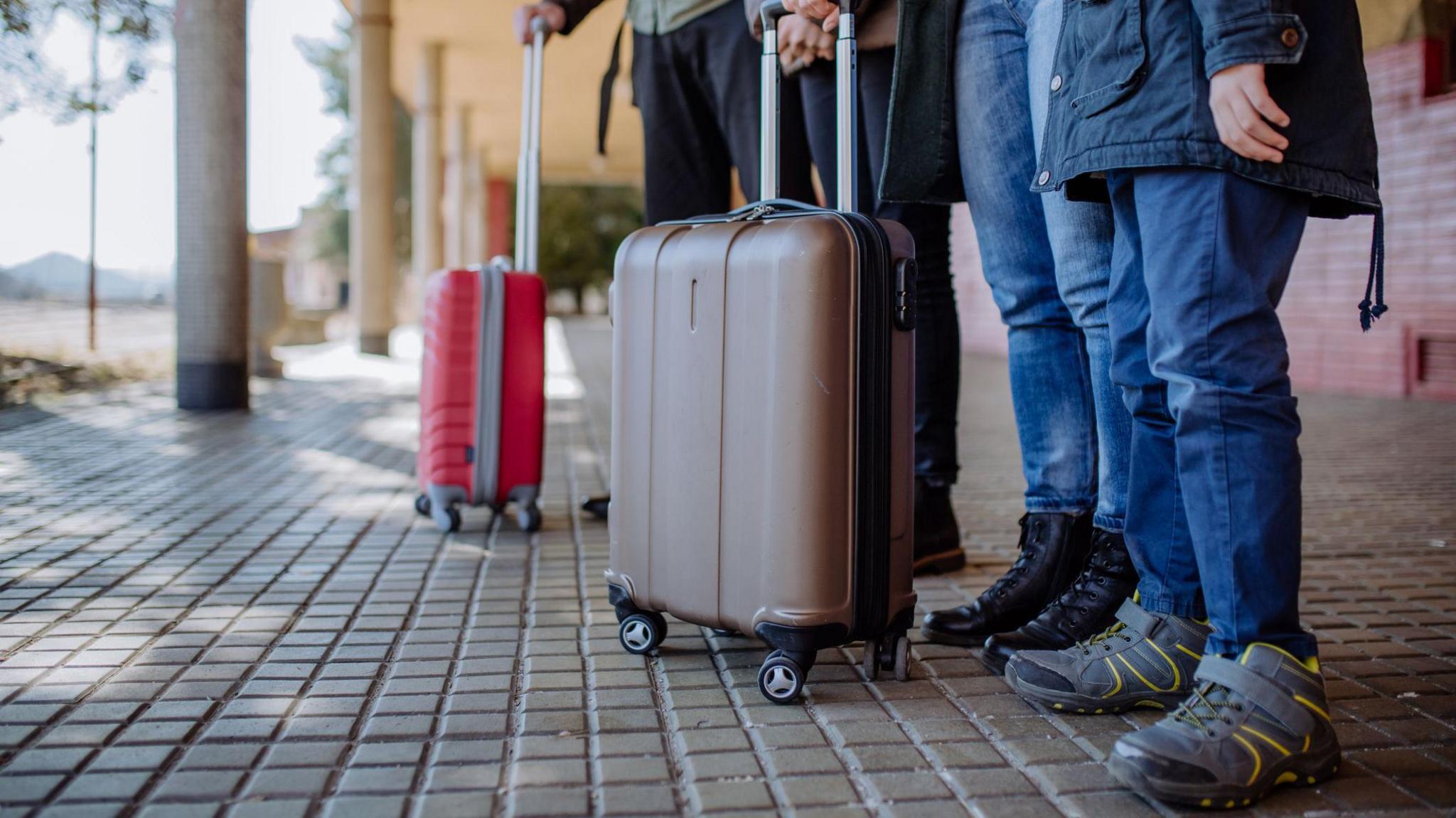 Four people, pictured from the waist down, standing behind two suitcases 