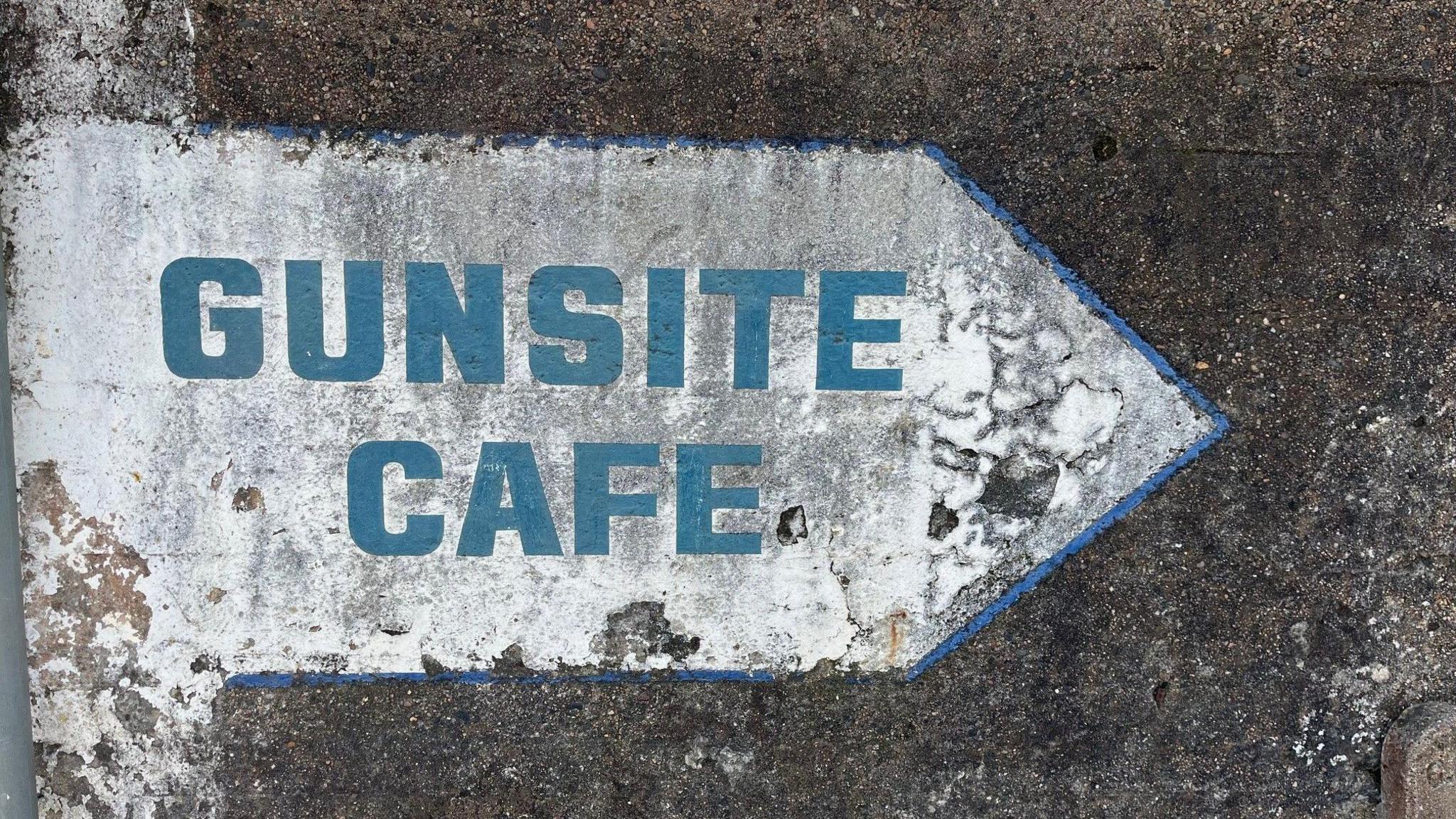 The words 'GUNSITE CAFE' on a sign which is shaped like an arrow on a dark grey wall.