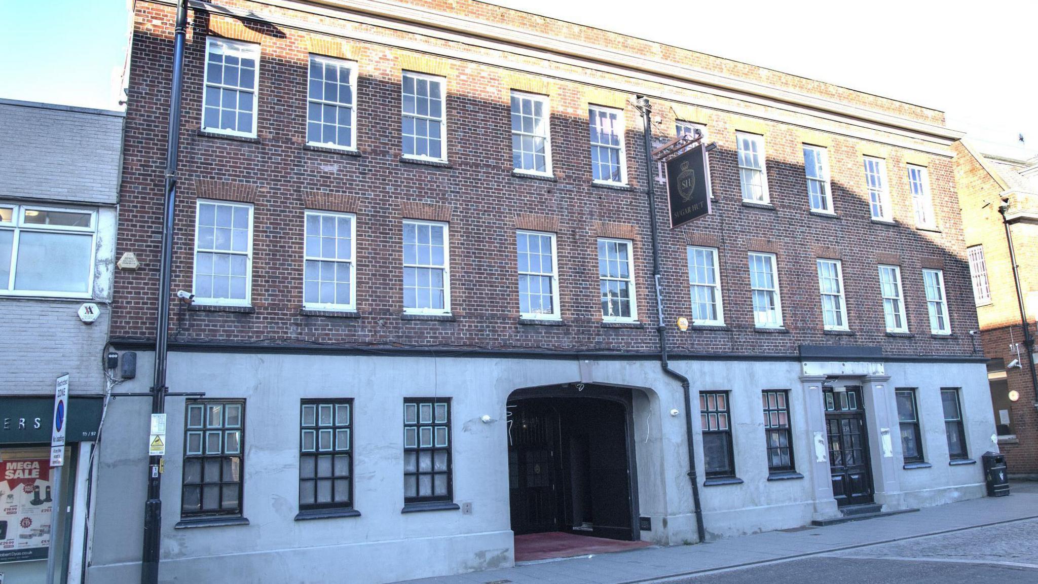 Sugar Hut, which is a three-storey building. The first and second floor have a brick facade, while the bottom one is made of white stone. All floors have many windows. There is an archway leading to the venue's main doors, which has a small red carpet in front of it. The venue is situated on a high street and pictured here on a sunny day.