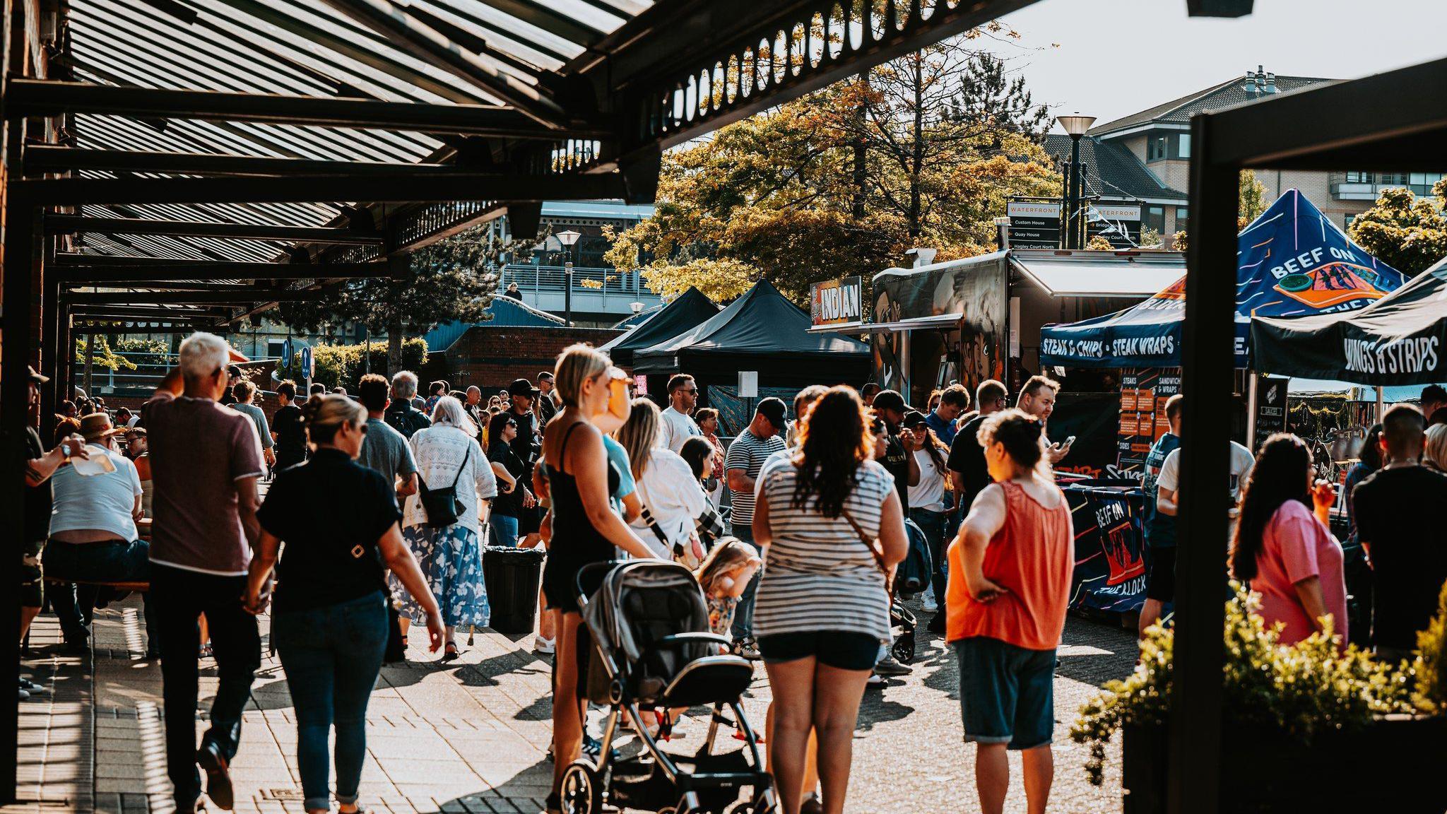 Groups of people walk among food stalls at the venue. It is a sunny day and the people are wearing summer clothes. There is canopy above them which has slates and the sun coming through parts of it. The food stalls have pointed roofs, which look like tents. 