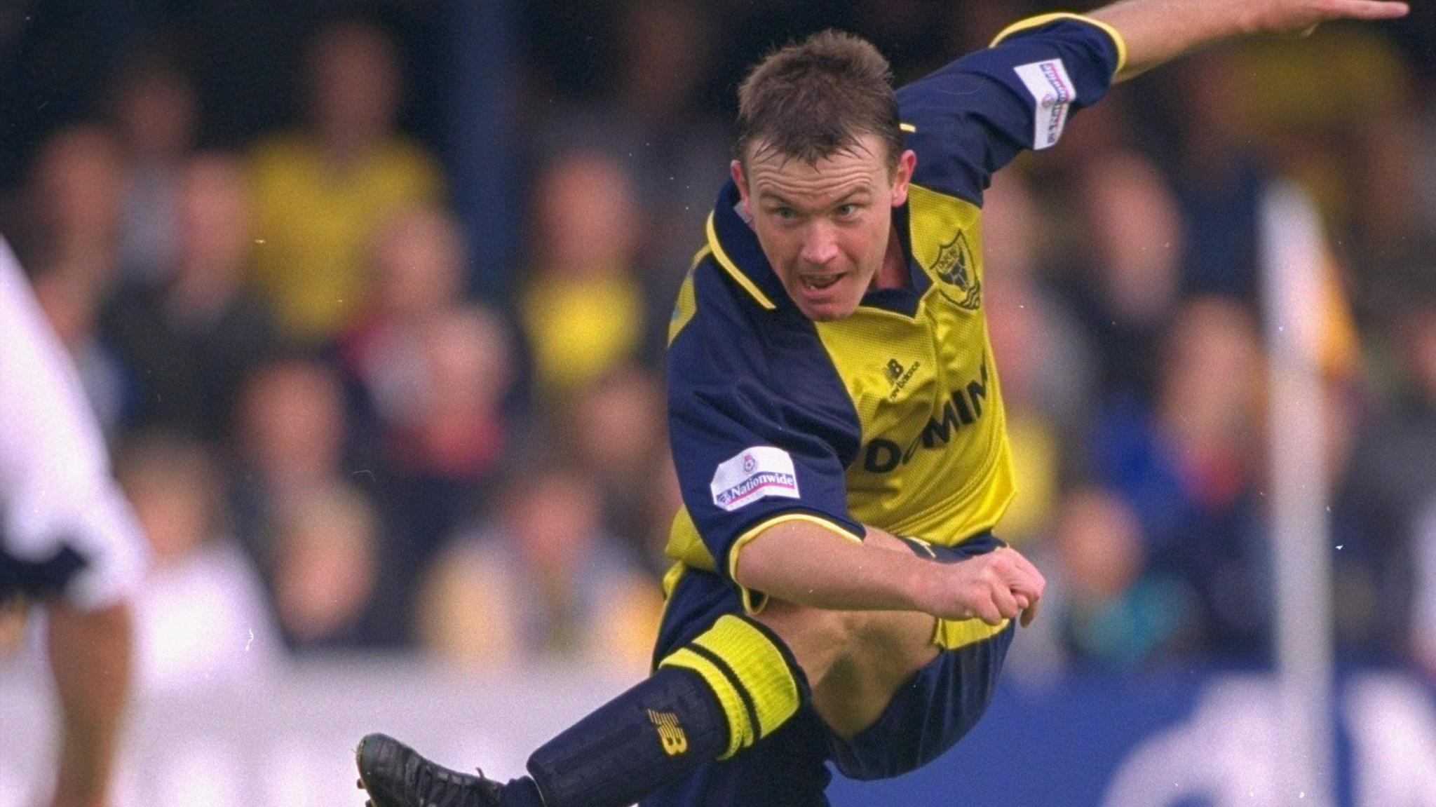 Joey Beauchamp of Oxford United in action. He has short brown hair and is wearing a yellow and blue Oxford jersey.