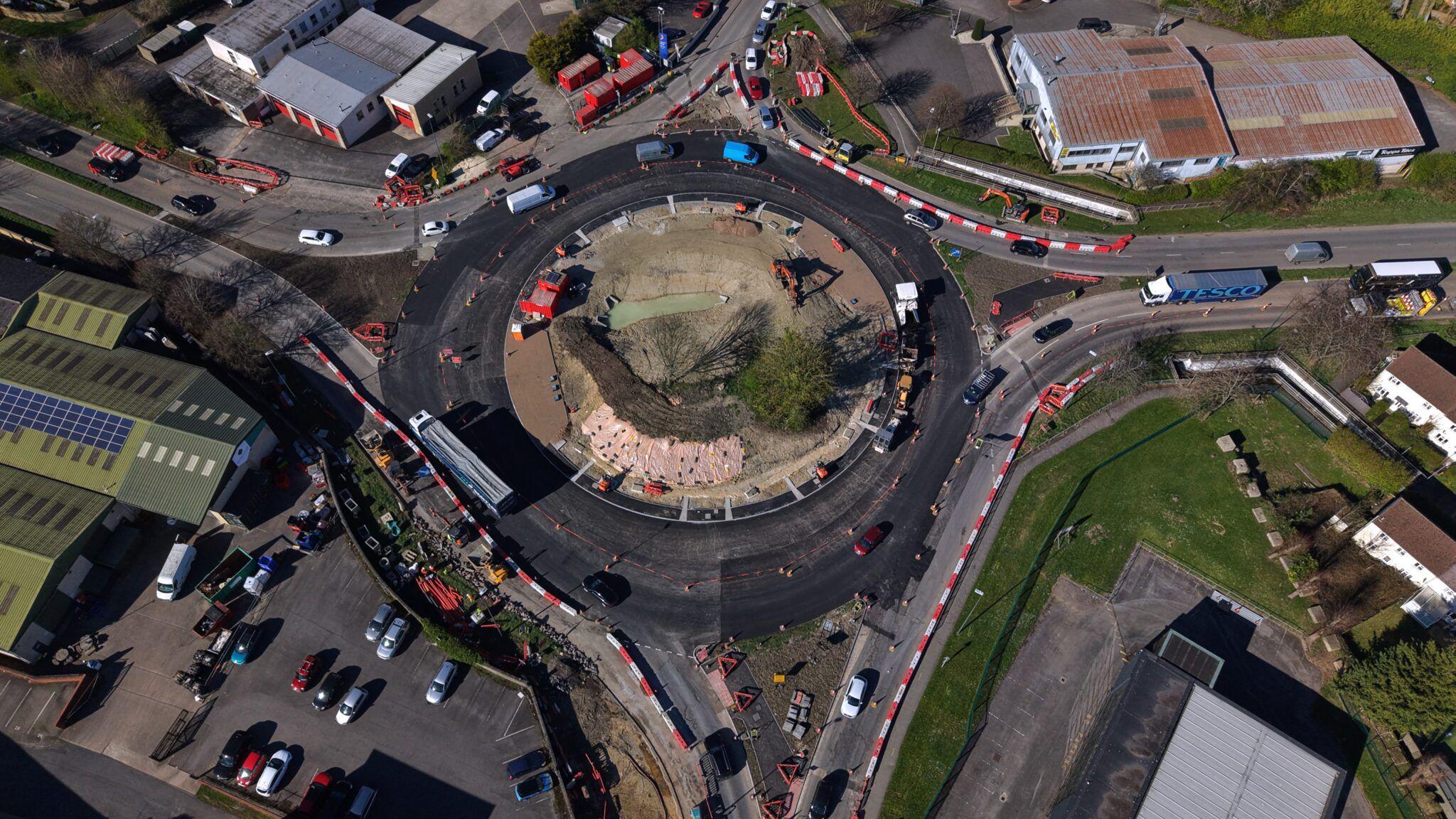 Drone image taken above a large roundabout where roadworks are taking place. There are four exits. Vehicles can be seen driving around the roundabout in lanes denoted by orange cones. The middle of the roundabout has been dug up and there are construction vehicles on it. An industrial and business area surrounds the roundabout.