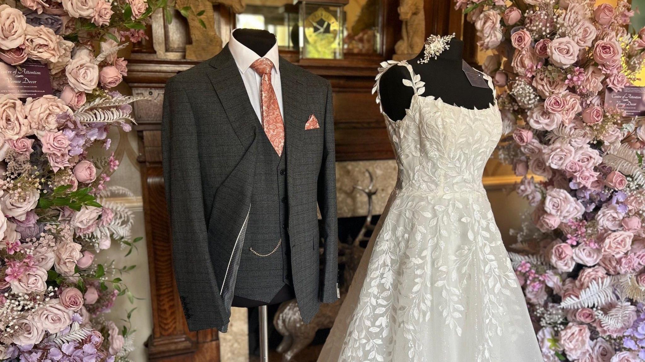 Dau fodel ochr yn ochr wedi eu gwisgo mewn dillad priodasol. Ar y chwith mae siaced smart a gwasgod lwyd, crys gwyn a thei batrymog binc. Ar y dde mae ffrog briodas ifori di-lewys. Mae blodau ffug pinc a phorffor golau wedi eu gosod bob ochr.