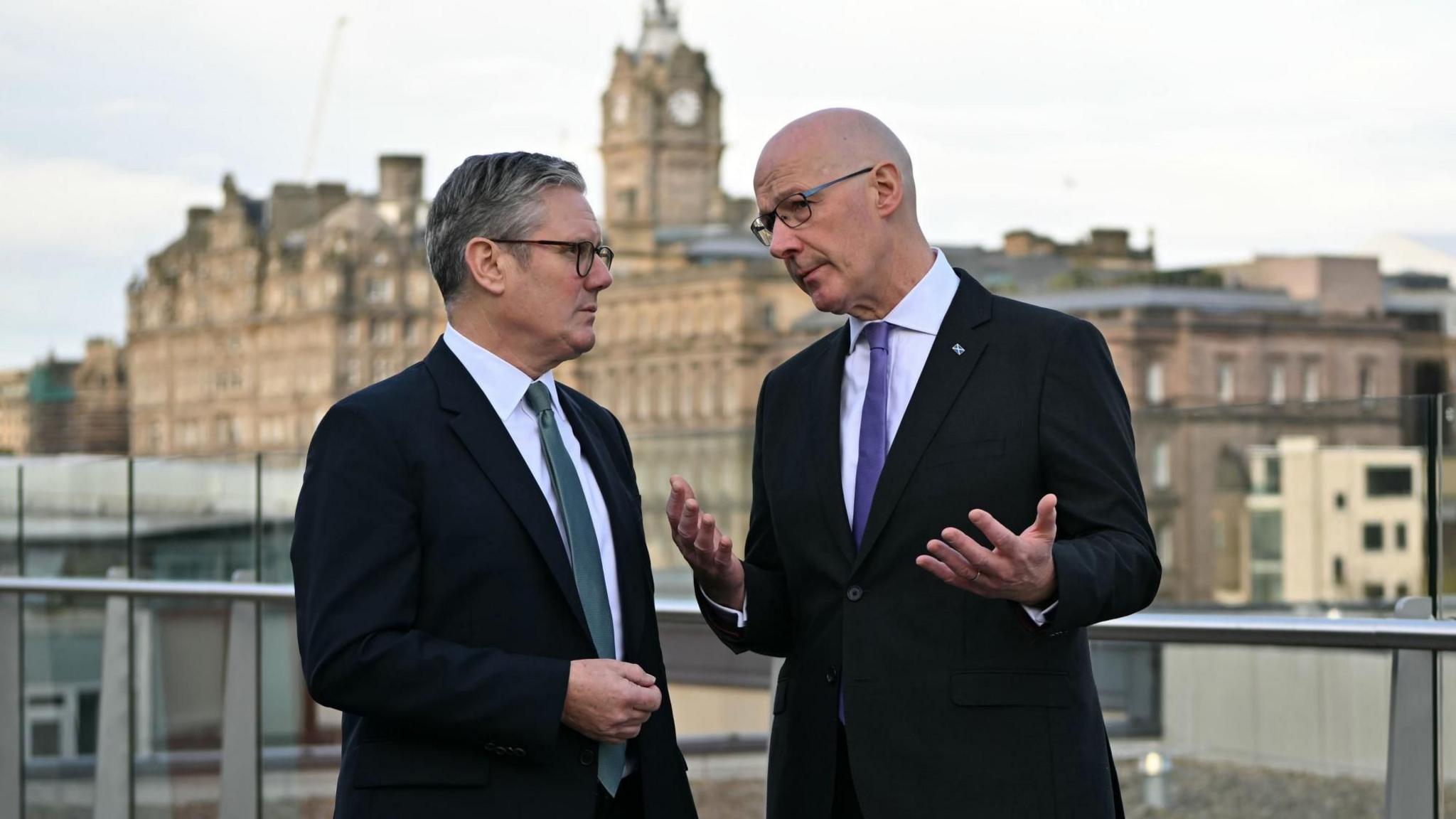 Sir Keir Starmer and John Swinney talking to each other with Edinburgh in the background. Both men are wearing dark suits, white shirts and ties.