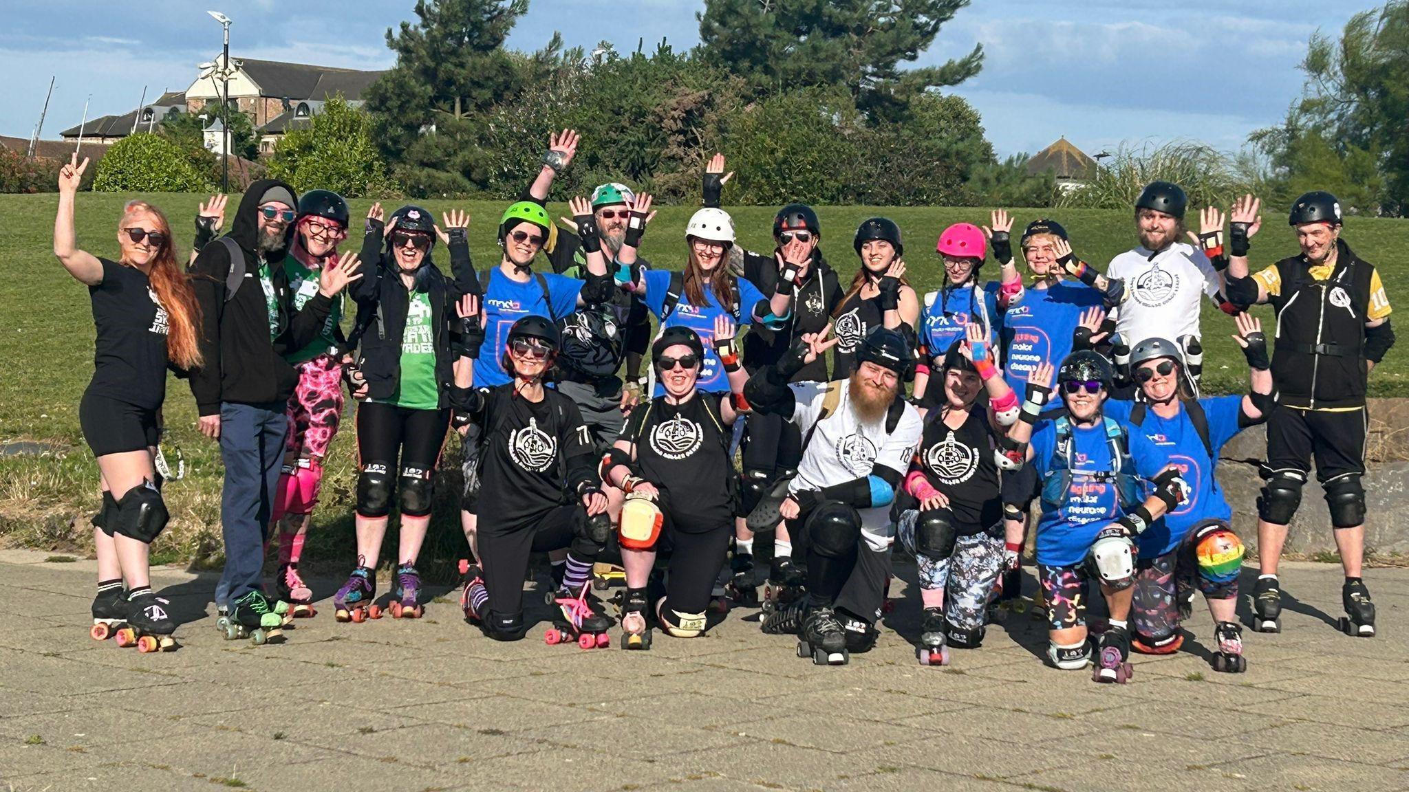 A large group of skaters waving all wearing colourful clothes and helmets.