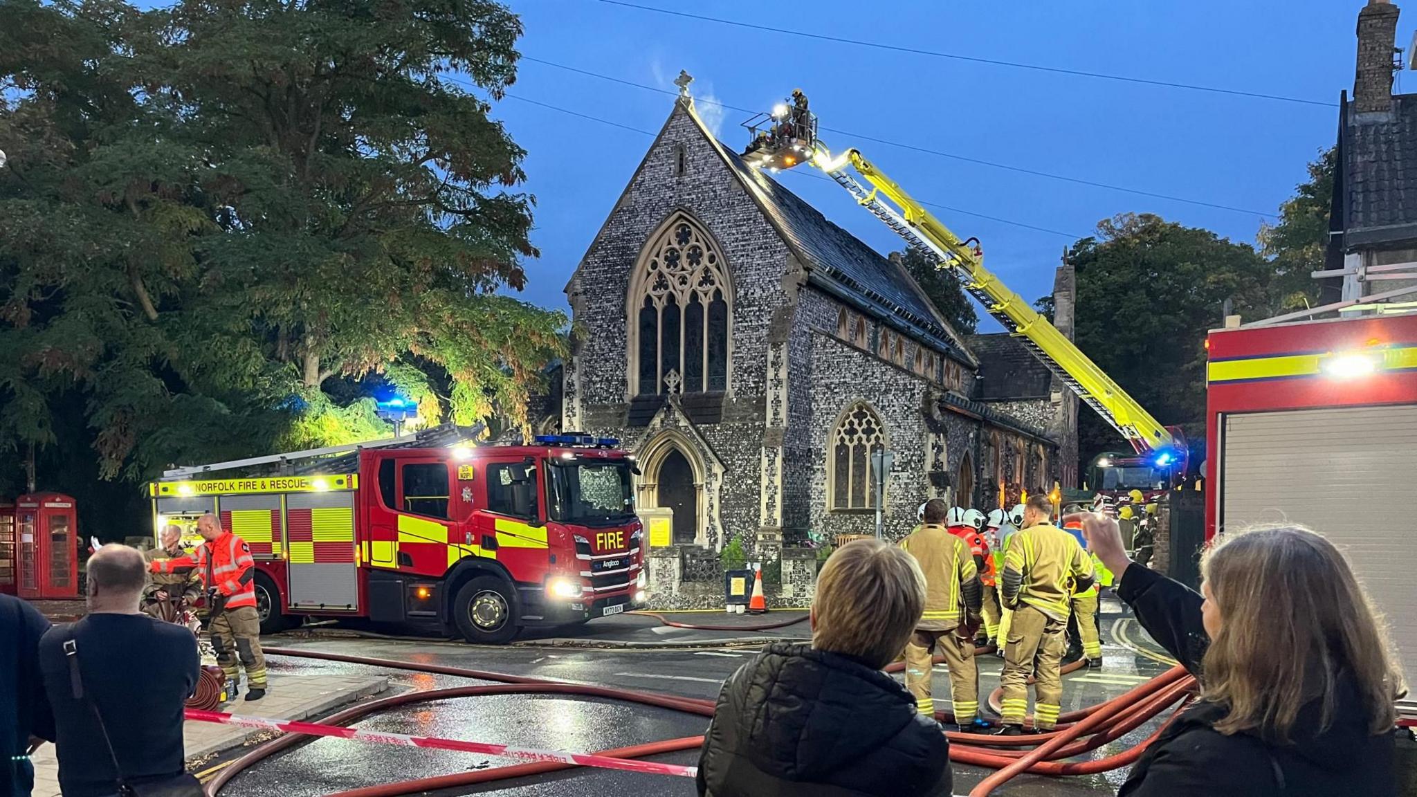 Firefighters on a ladder just above the roof of a church, just behind its pointed gable end. A small amount of smoke is behind the tip of the gable end, which has a cross at its apex.  Fire engines and passers by are in the street. There are red hoses across the road.
