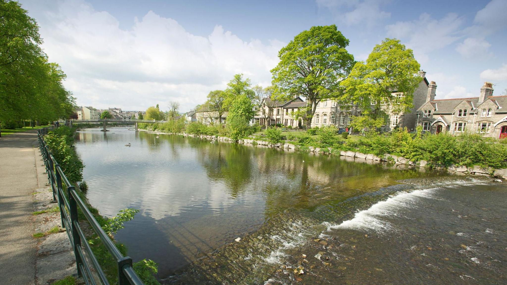 River Kent, seen from Abbot Hall Park in Kendal, Cumbria, looking over riverside railings and across to some pretty stone houses and trees on the other riverbank.