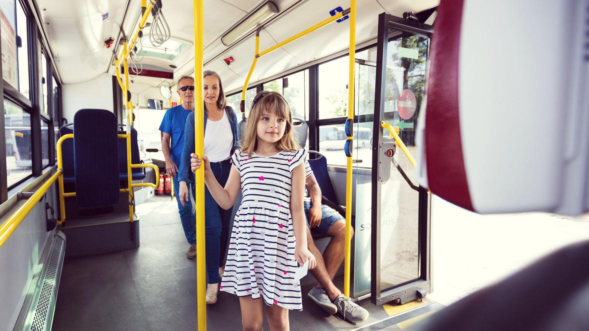 A girl in a white and black striped dress holds on to a yellow handrail on a bus.