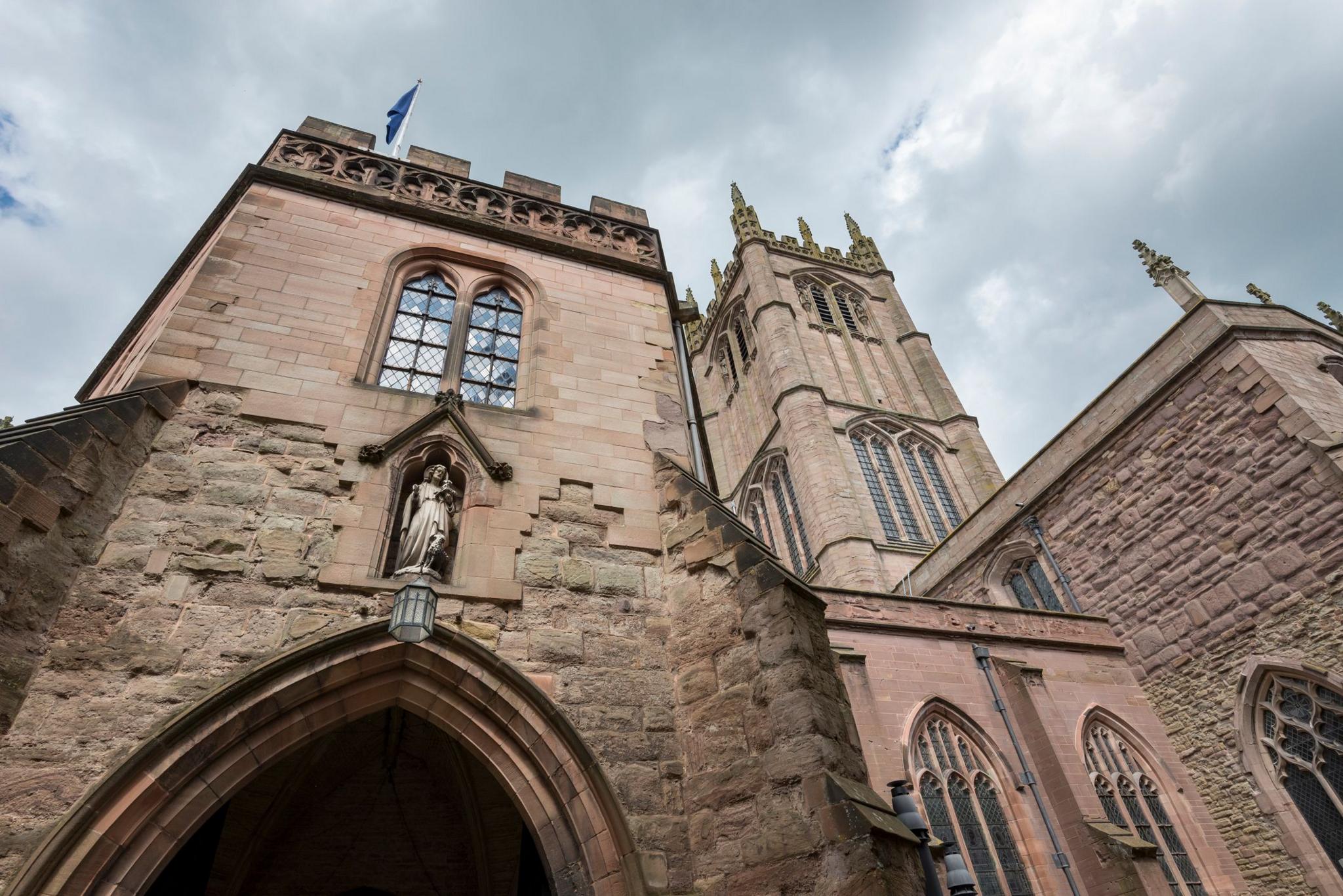 A shot of a large stone church building, taken from low level looking up at the tower and other parts of the church. A blue flag flies from one crenellated roof. 