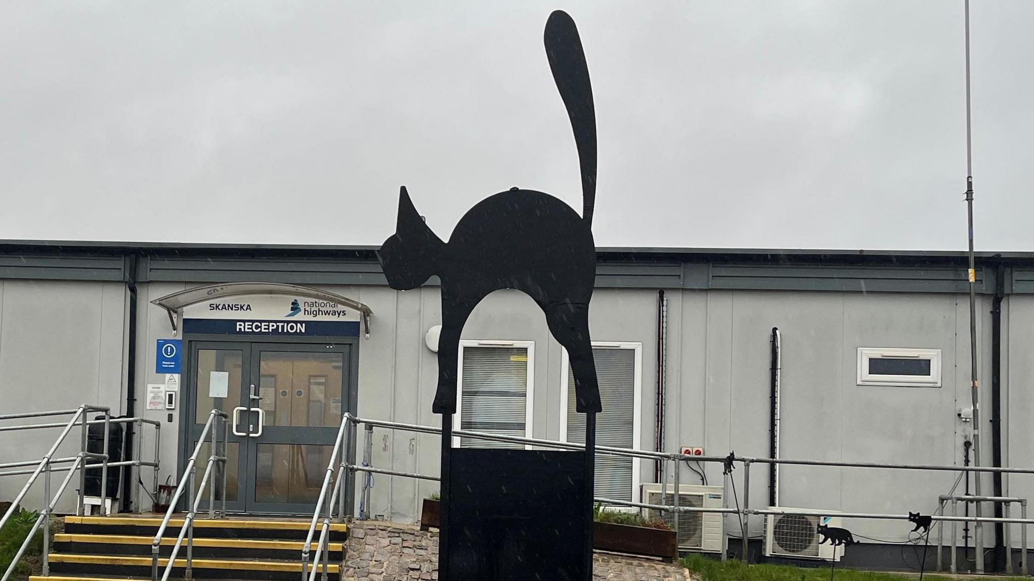 A statue of a black cat with its tail in the air outside a National Highways compound.