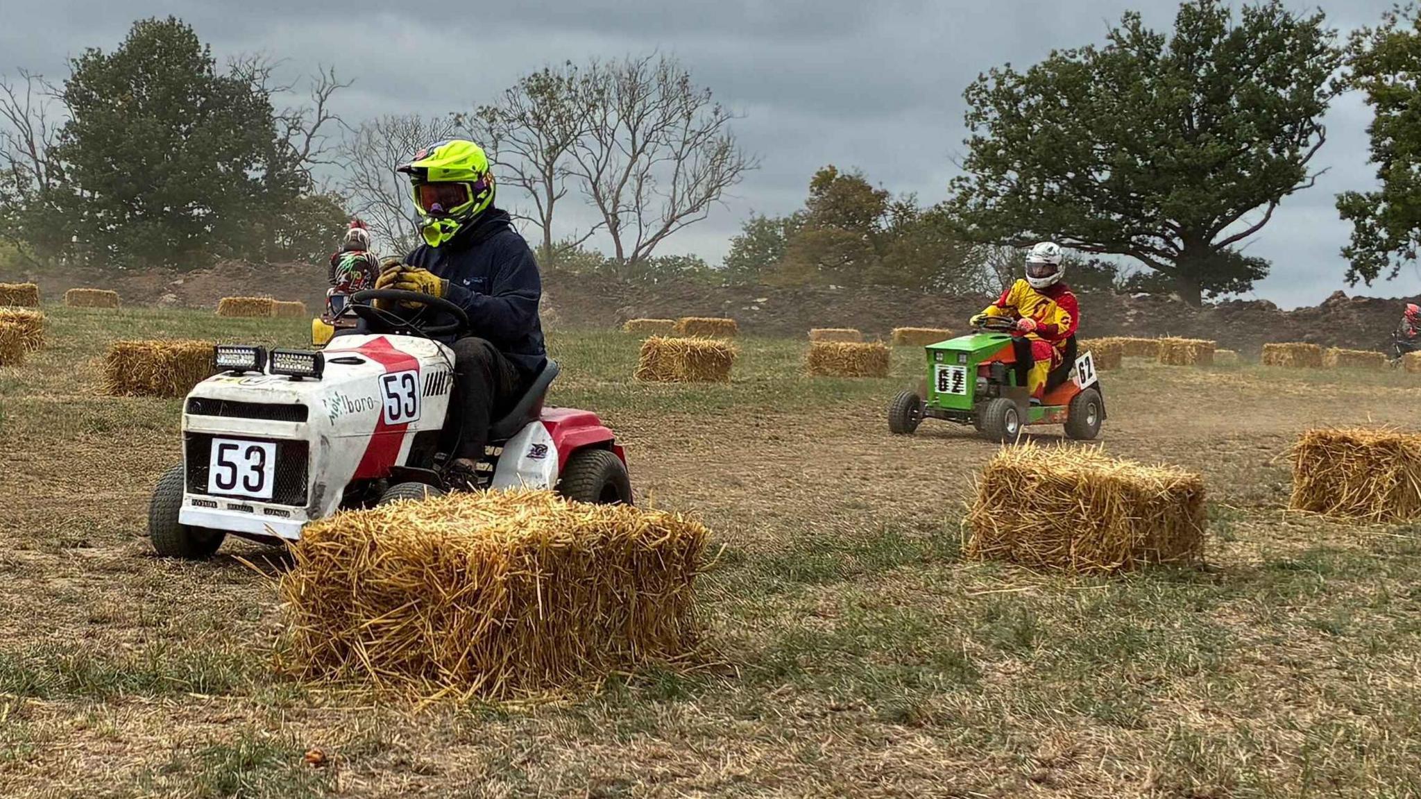 Two men on lawn mowers with racing colours and numbers racing down a a dirt track. 