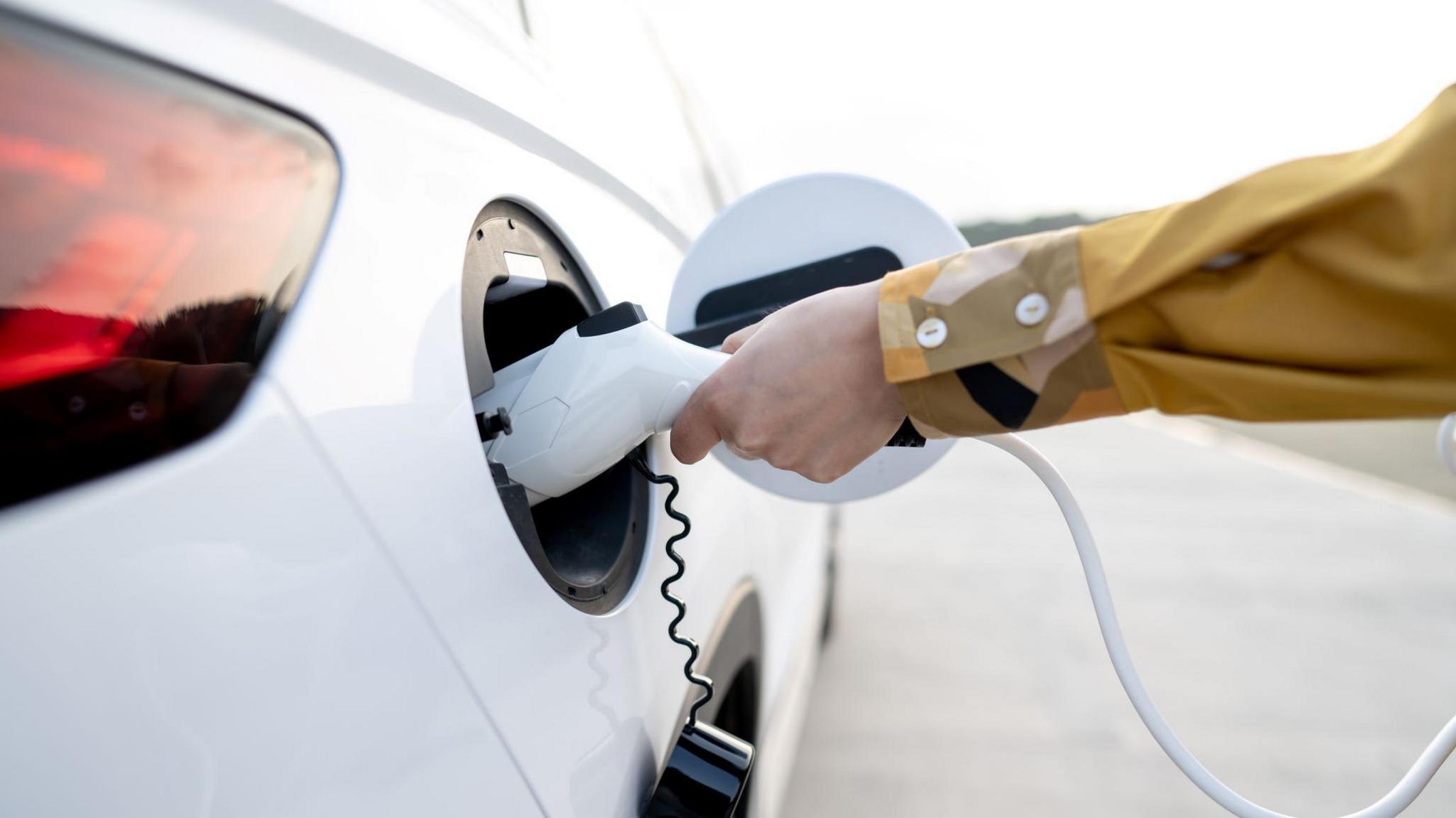 A hand and arm of a person wearing a mustard-coloured shirt connects an EV charger to a white car.