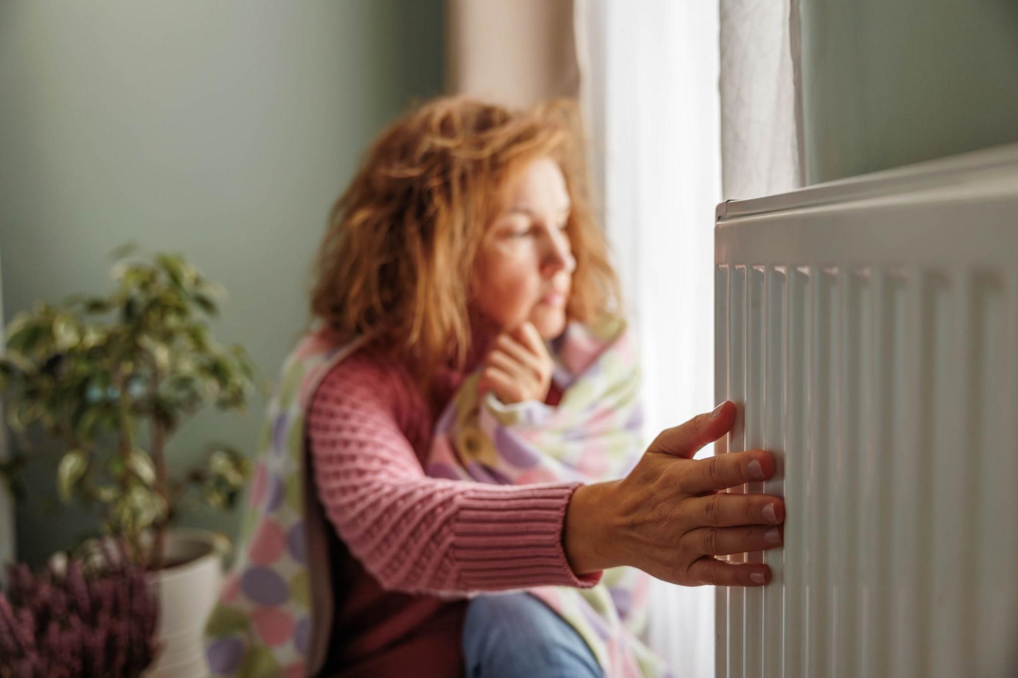 Woman wearing a jumper and wrapped in a blanket puts her hand on a radiator. A plant is in the background.