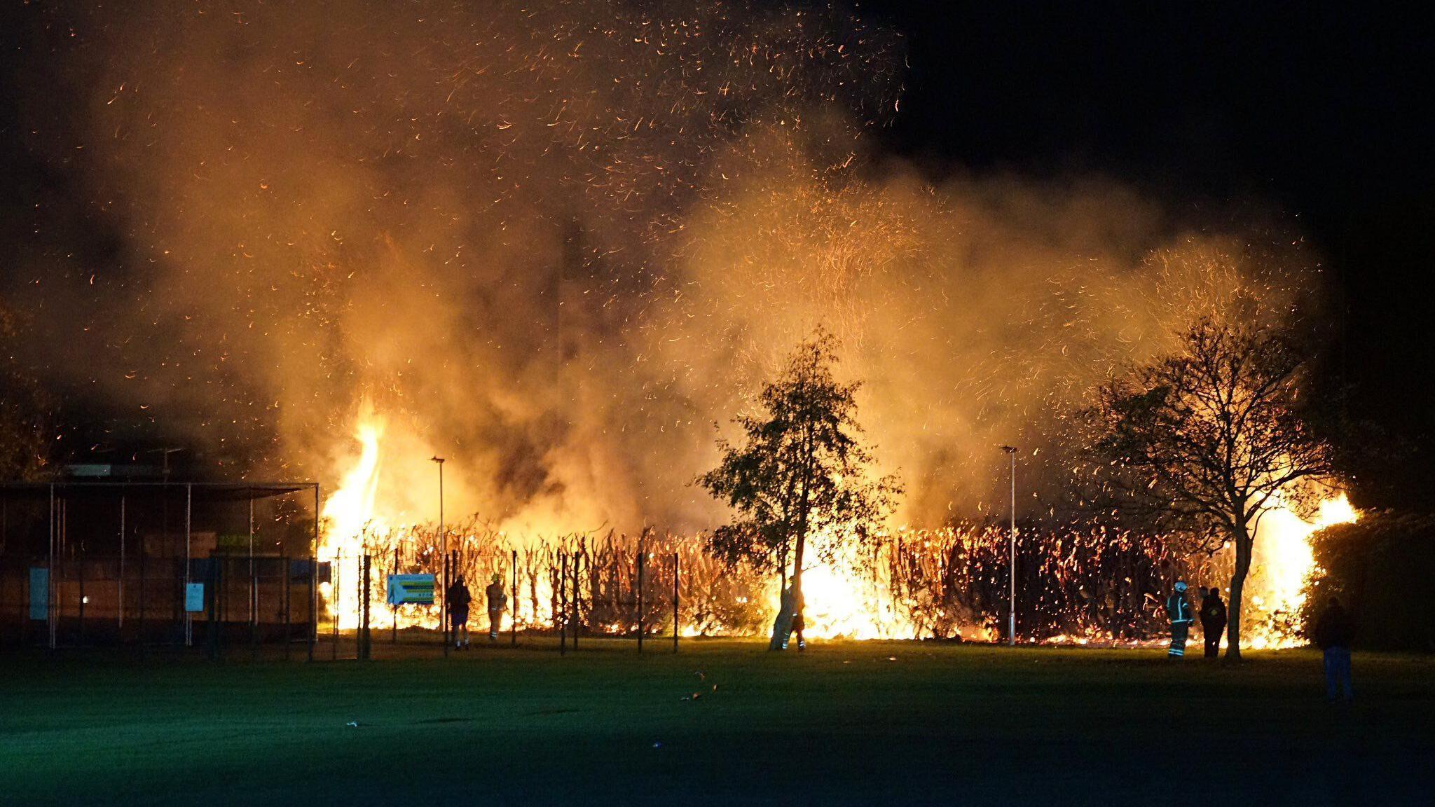 Large flames coming from hedges and trees surrounding football pitches. The sky is dark and firefighters and members of the public are standing nearby.