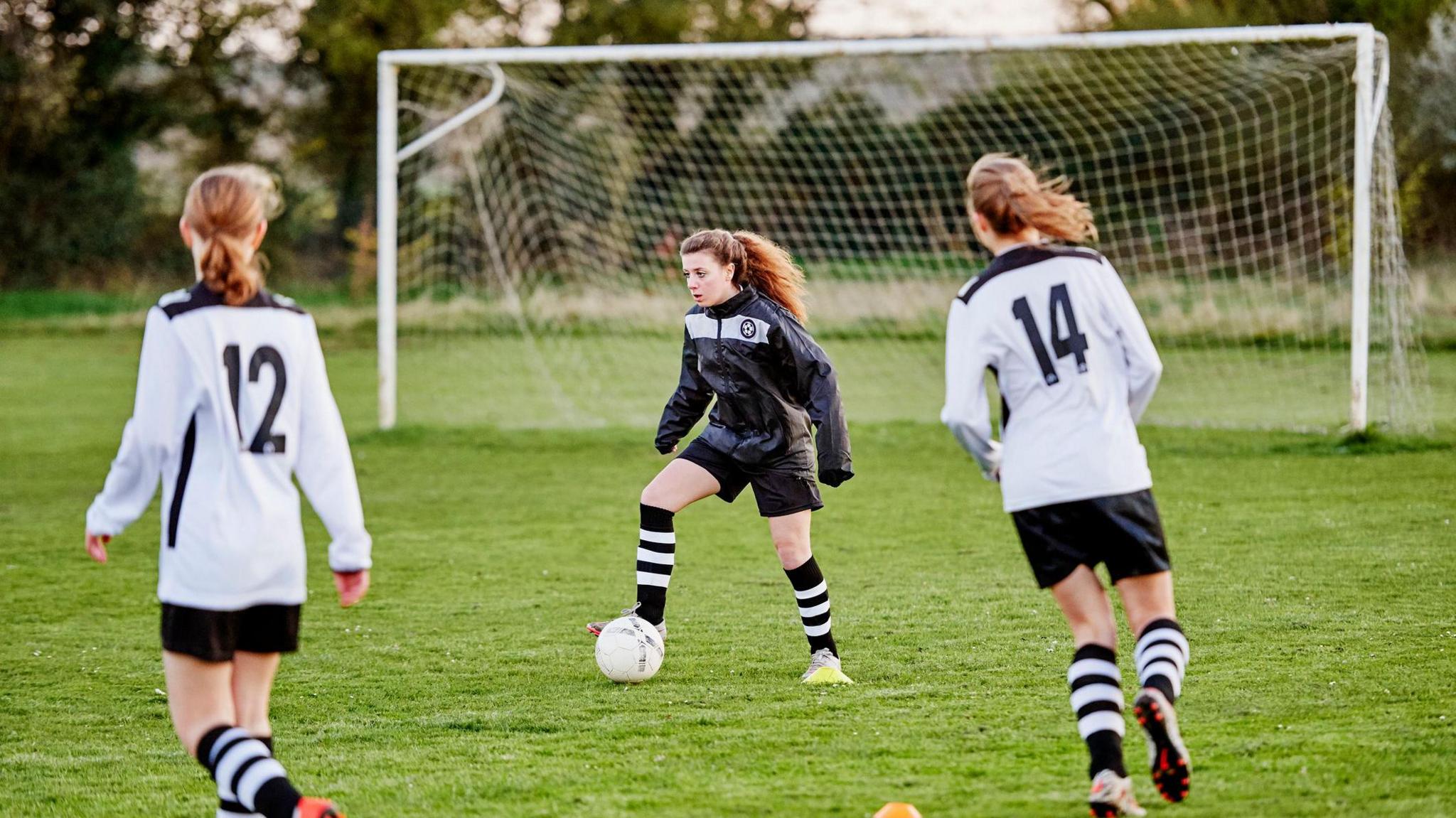 Three teenage girls playing football. The player in the middle is wearing a black kit, including a waterproof jacket and is dribbling the football. Two players on the opposing team, wearing white kits, are heading towards her on either side.