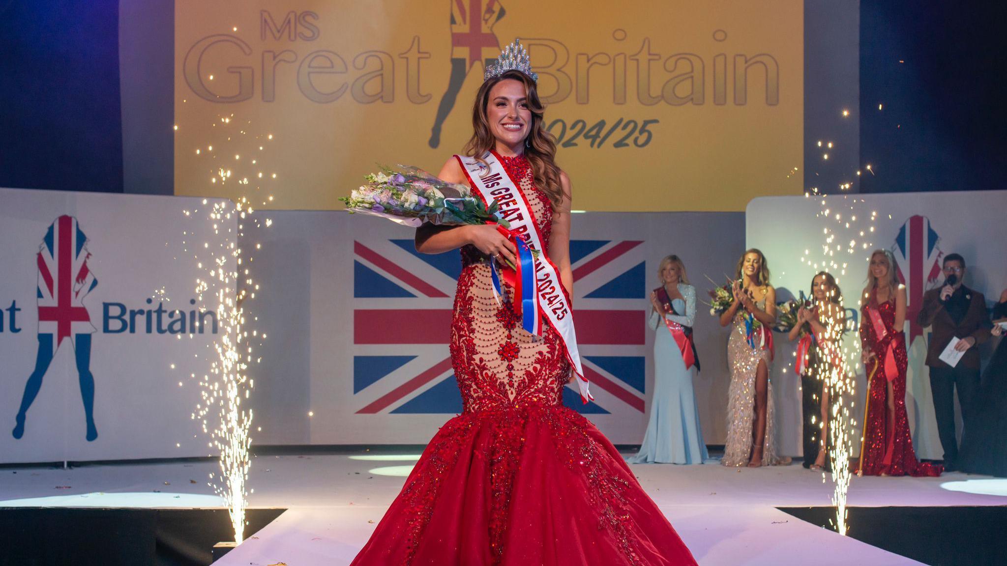 Emma on stage is a red ballroom dress, with a MS GB 2025 sash in white and a diamond tiara. she's holding a bouquet of flowers. There are fireworks going off and you can see other contestants in their gowns in the background 