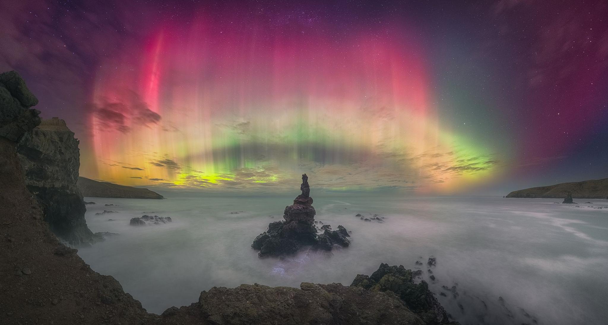 The Northern Lights in pink orange and yellow dance over the sky and ocean.