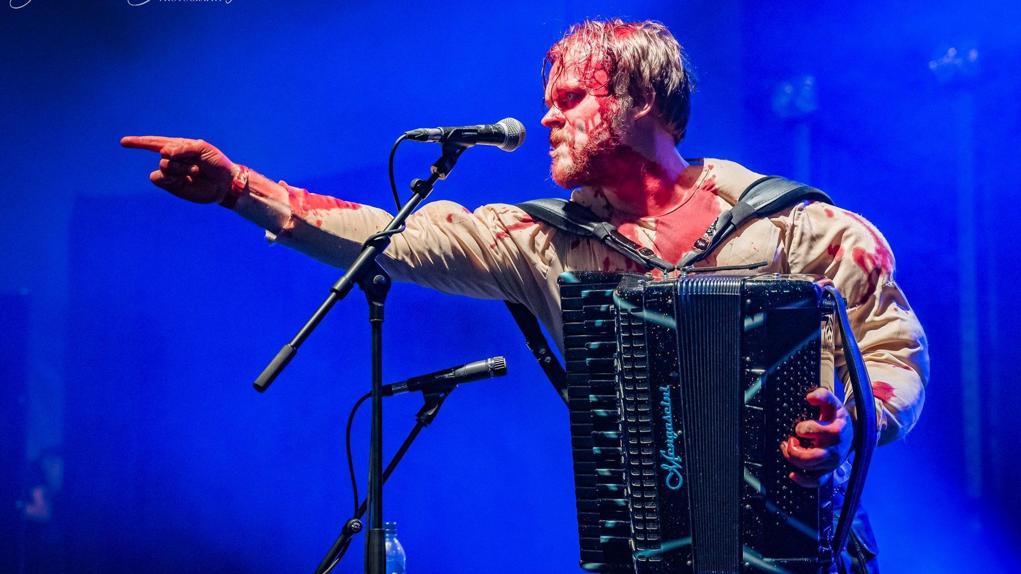A man holding an accordion, with fake blood/paint over him. He is standing at a microphone and pointing to something away offstage.