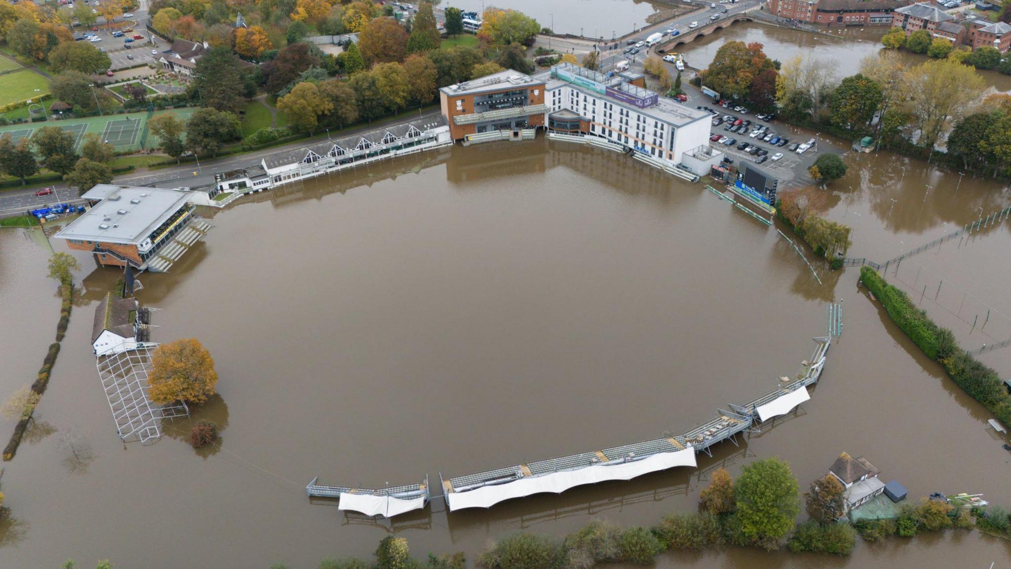 An aerial shot of New Road Cricket Ground in Worcester, submerged under brown floodwater.  The upper parts of stands and buildings can be seen around the water which covers a large area of the photograph.