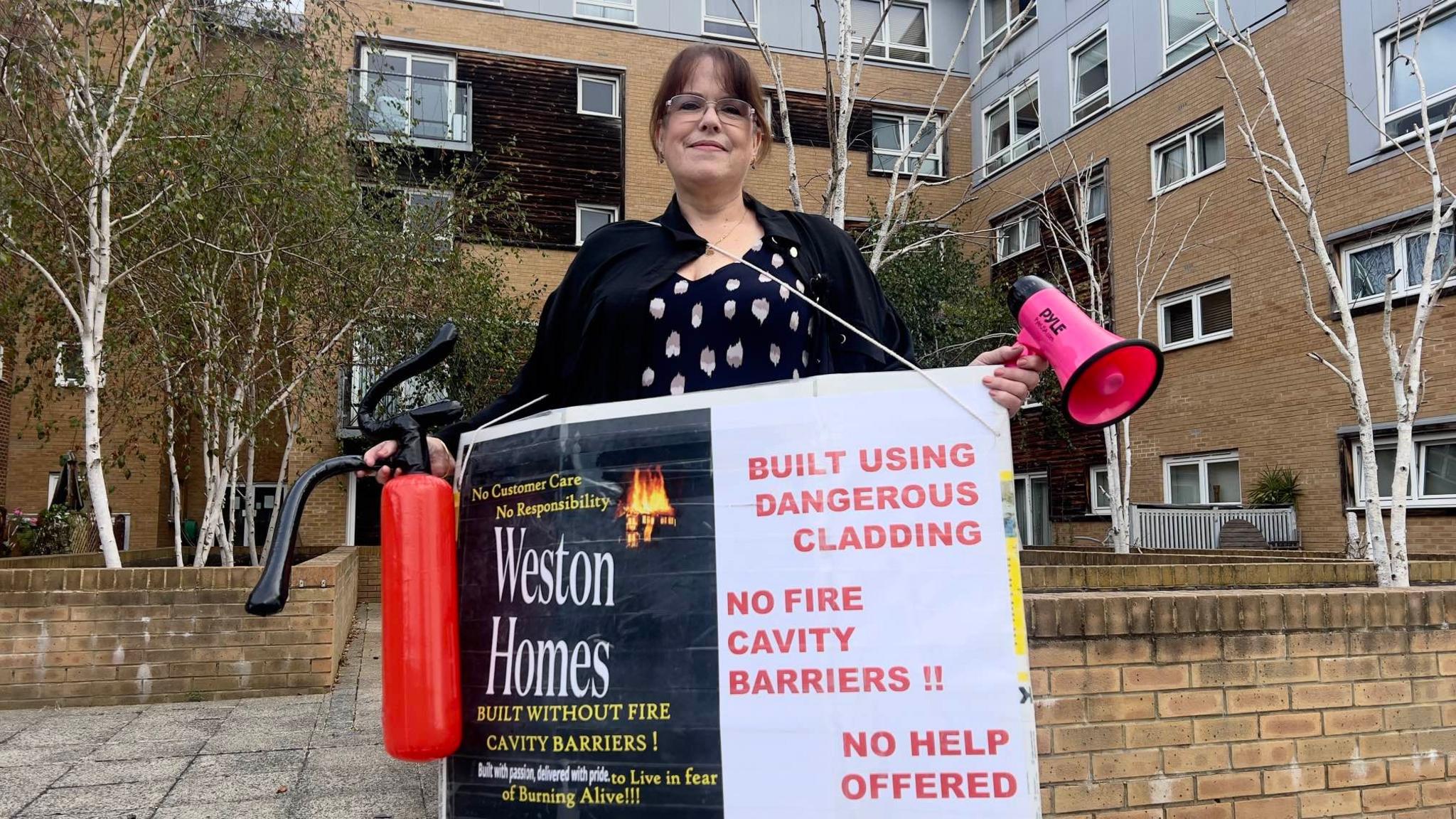 Jane Randle, who is holding a megaphone and a fire extinguisher. She is also holding a sign criticising Weston Homes, claiming it has offered no help to residents and built the flats using "dangerous cladding". Jane herself is wearing glasses.