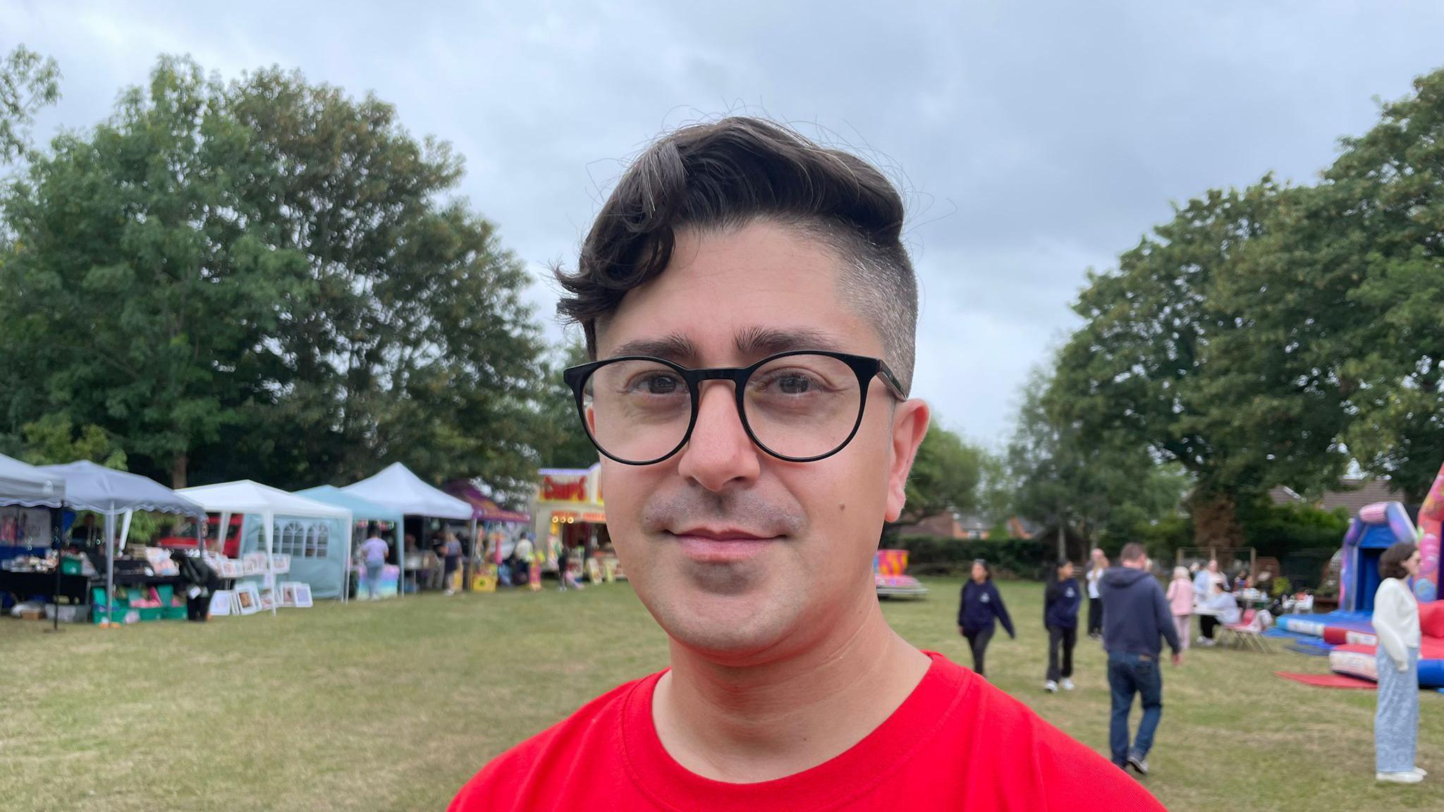 A man with short dark hair, Ish Shah, is wearing large black glasses and a red T-shirt and standing in a grassy field with large trees in the background. There are people near tents with various crafts and inflatable structures on the field.