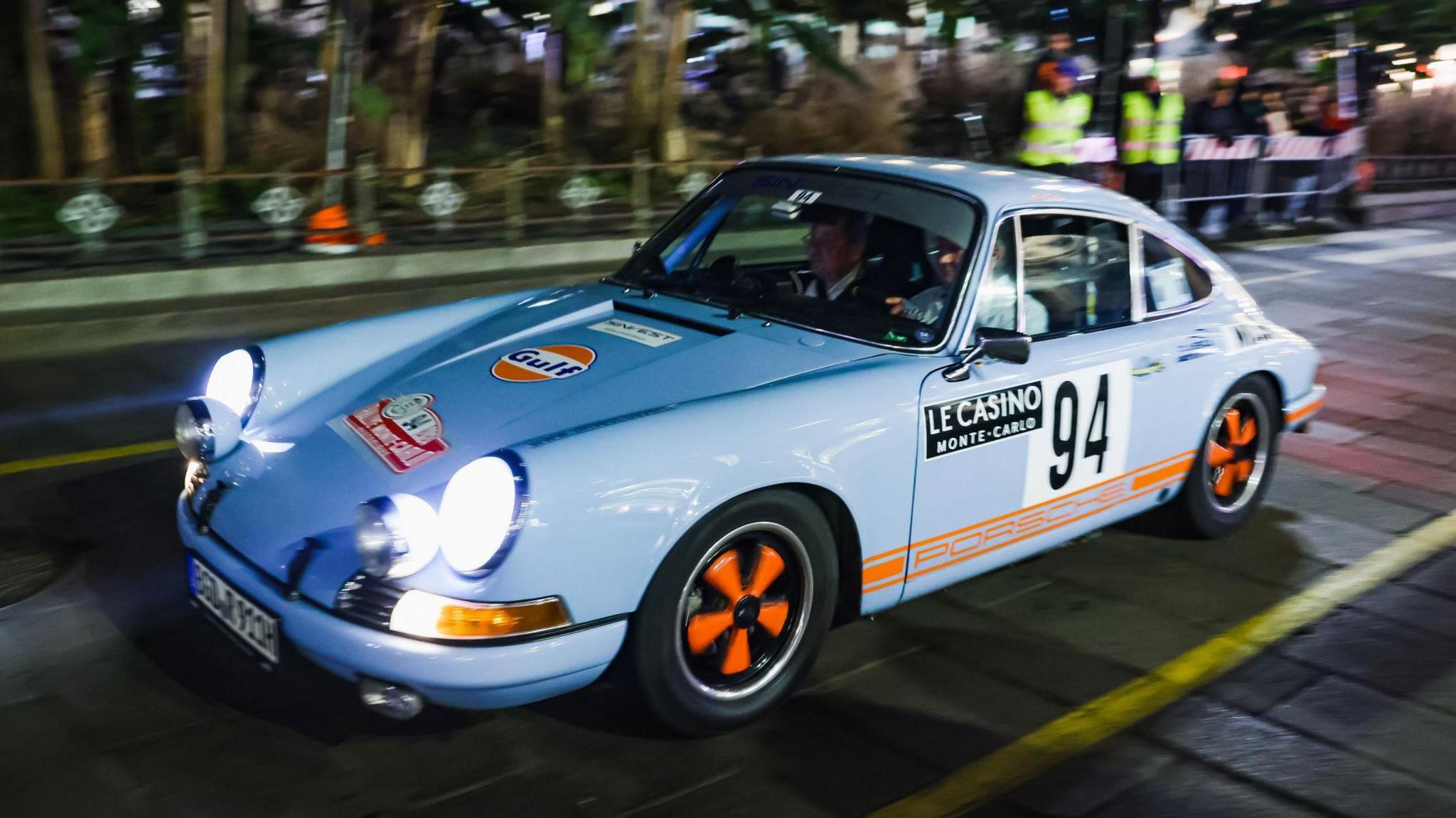 A blue Porsche is driven through a street in Milan during 2024's Monte Carlo Classique/Historique Rally. It is night-time and the car has its lights on.