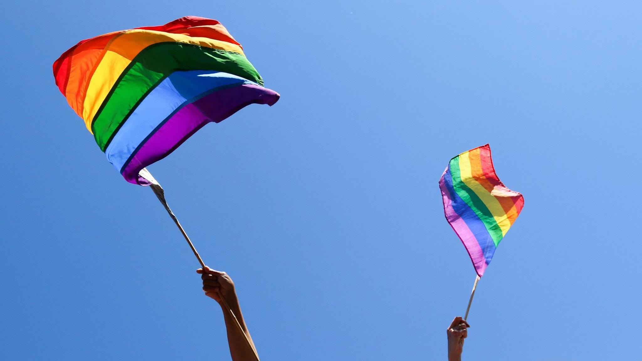 A photo of two hands waving pride flags against a blue, cloudless sky