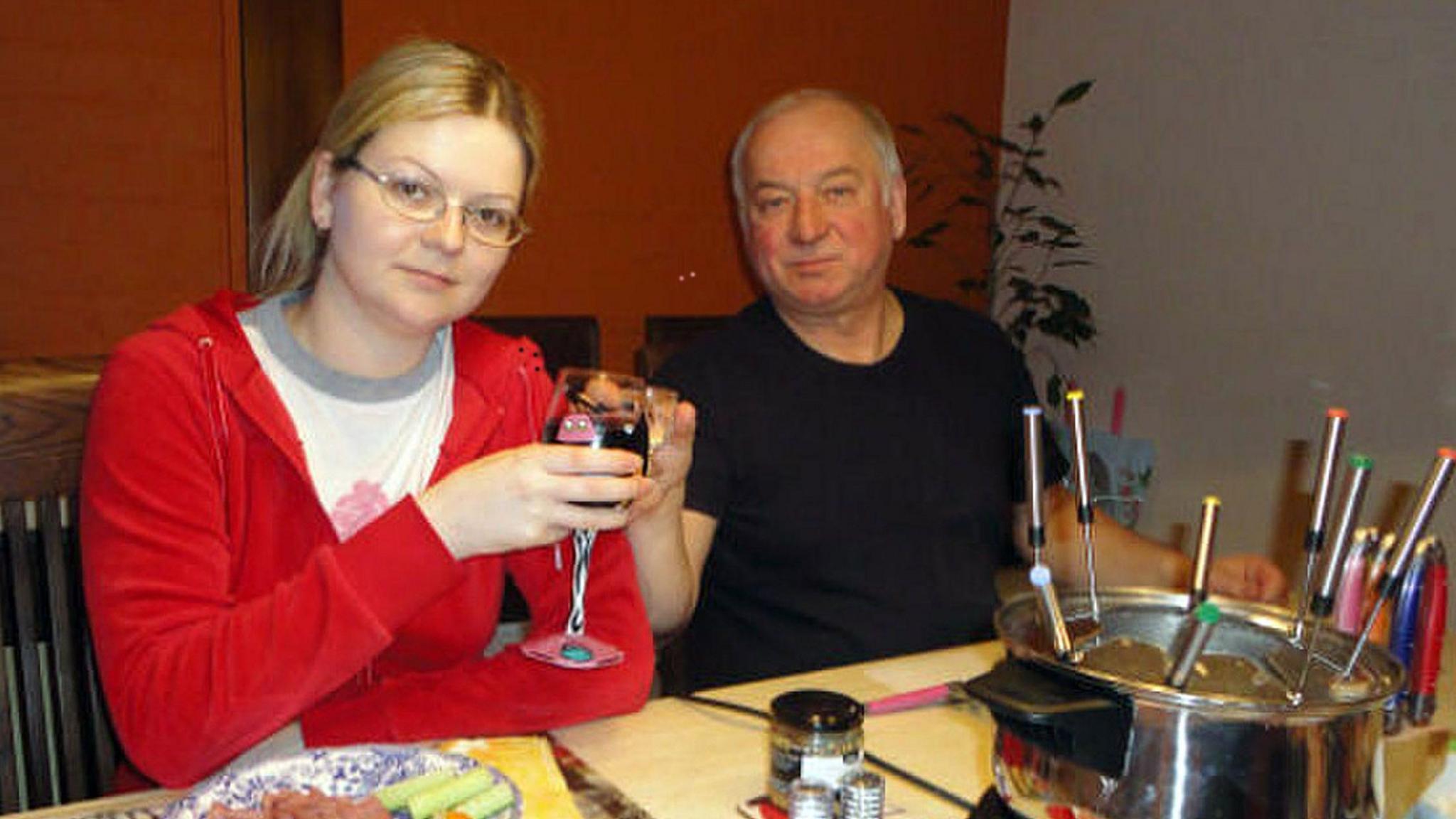 A woman in a red top clinks a glass of wine with an older man in a dark t-shirt at a table with a fondue set. 