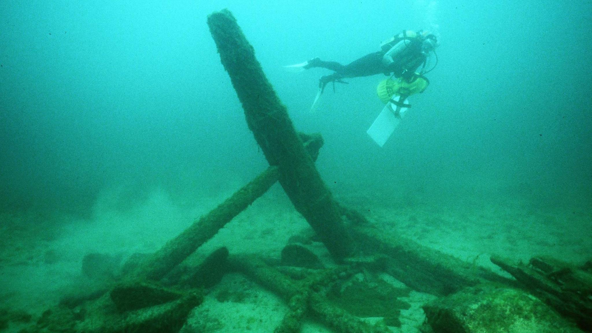 An underwater shipwreck, the water has shades of green and blue, a diver is swimming by what looks like an anchor