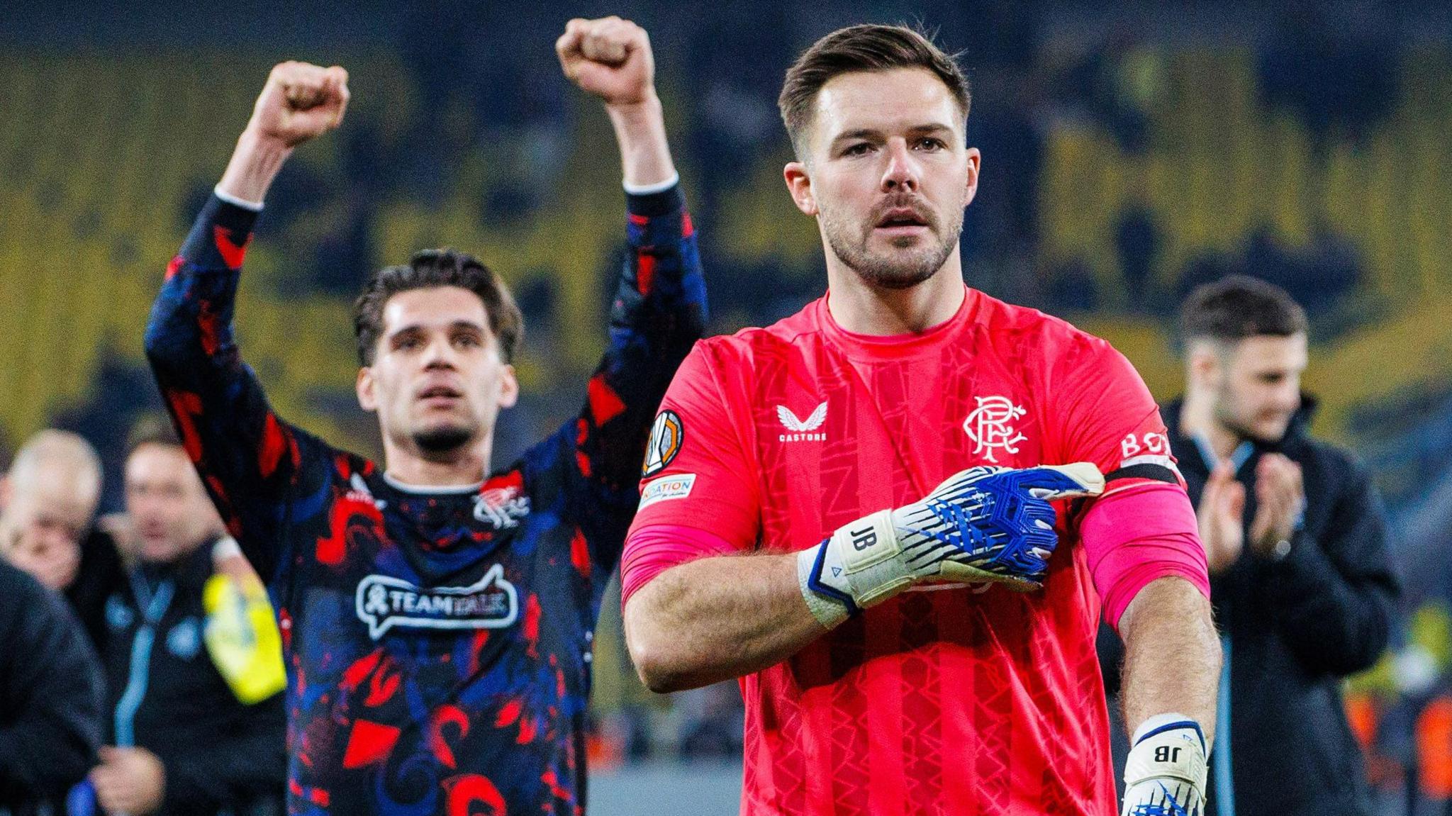 Jack Butland wearing a bright red shirt. He is pointing to the black armband on his left arm with a blue and white glove.