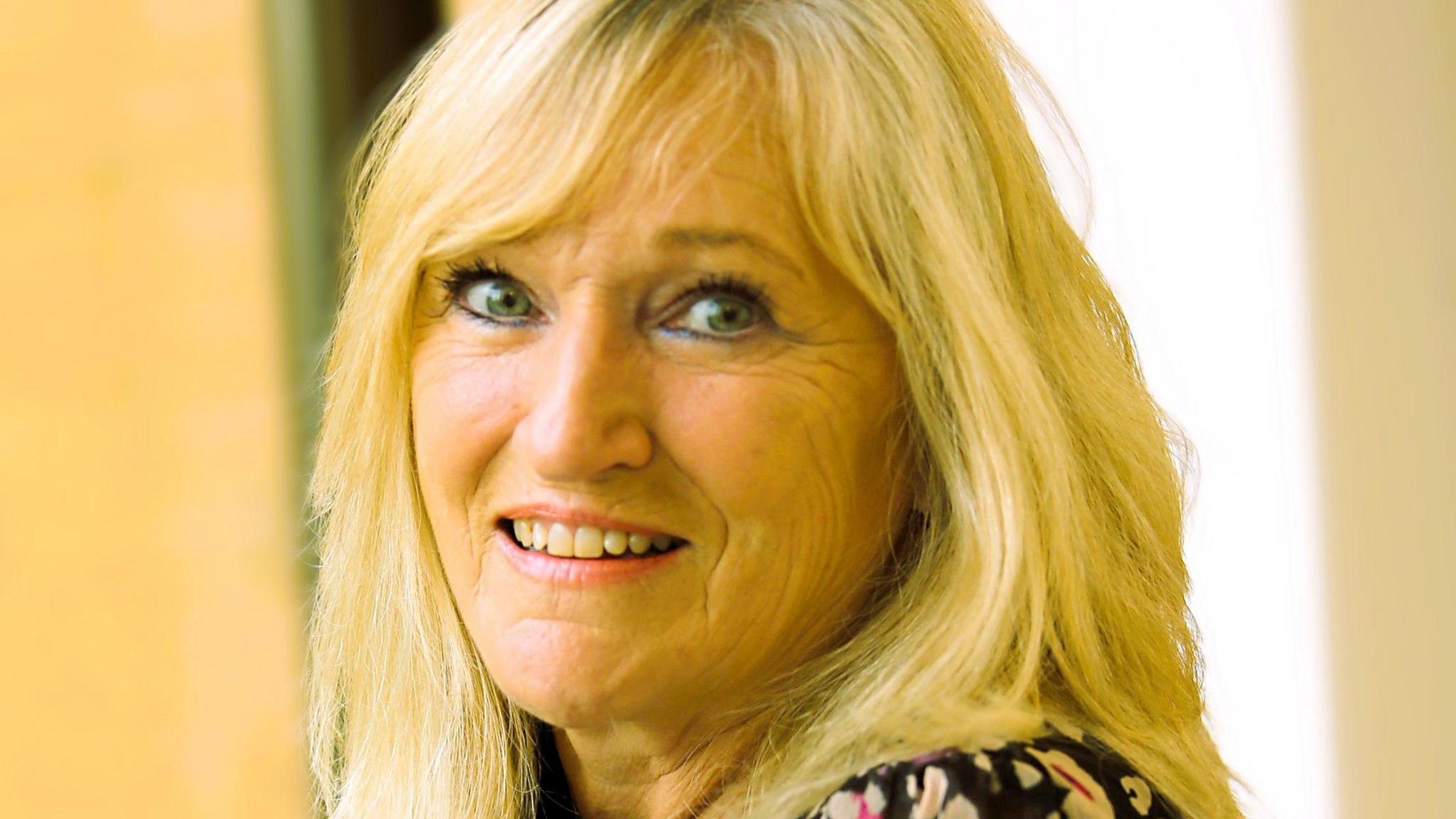 Karen Loweman has long blonde hair and is wearing a floral dress. She is smiling at the camera indoors.