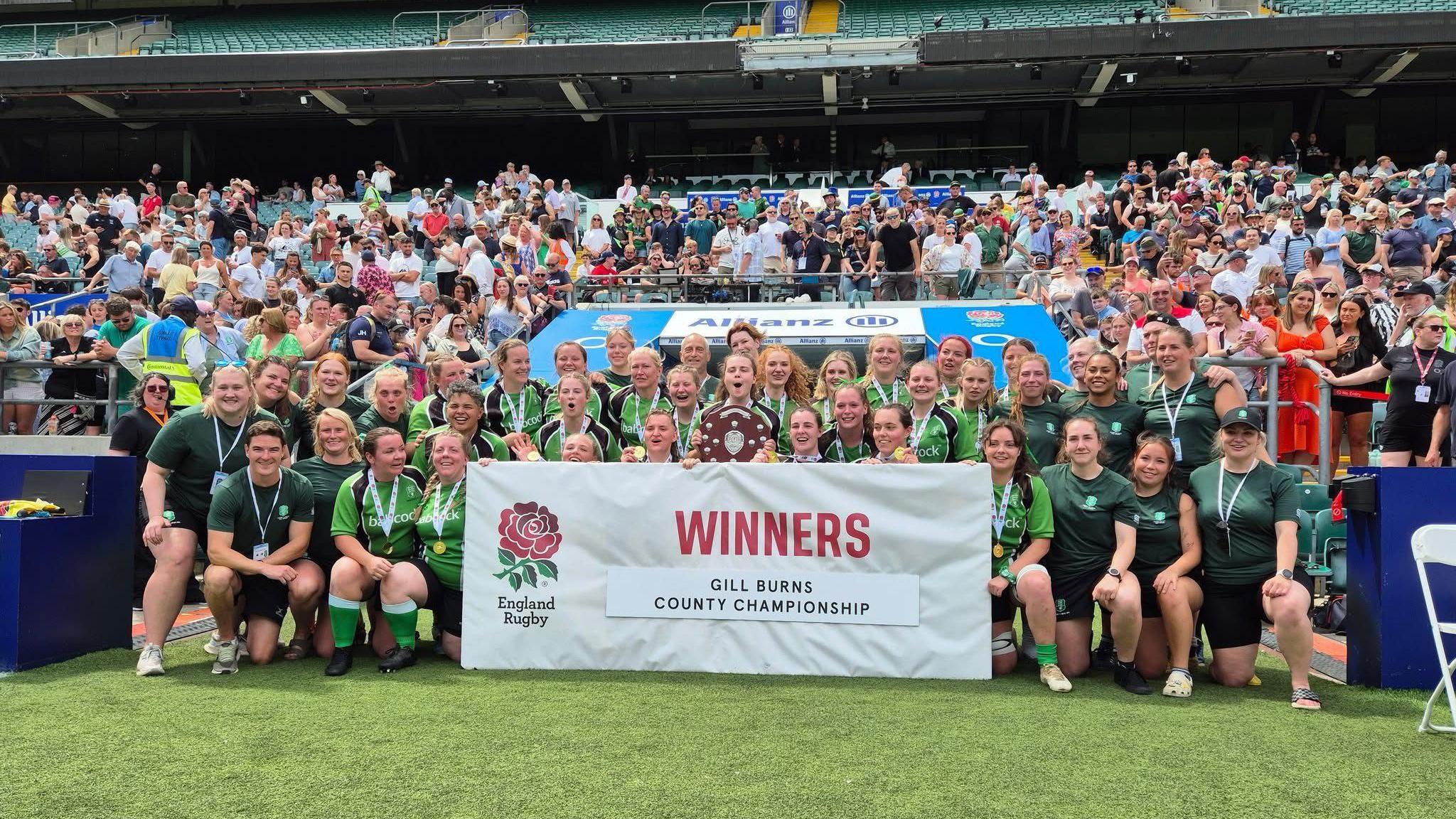 Women wearing green kits gathered around a white banner on the pitch, which reads "Winners - Gill Burns County Championship". The stand behind is busy with fans.