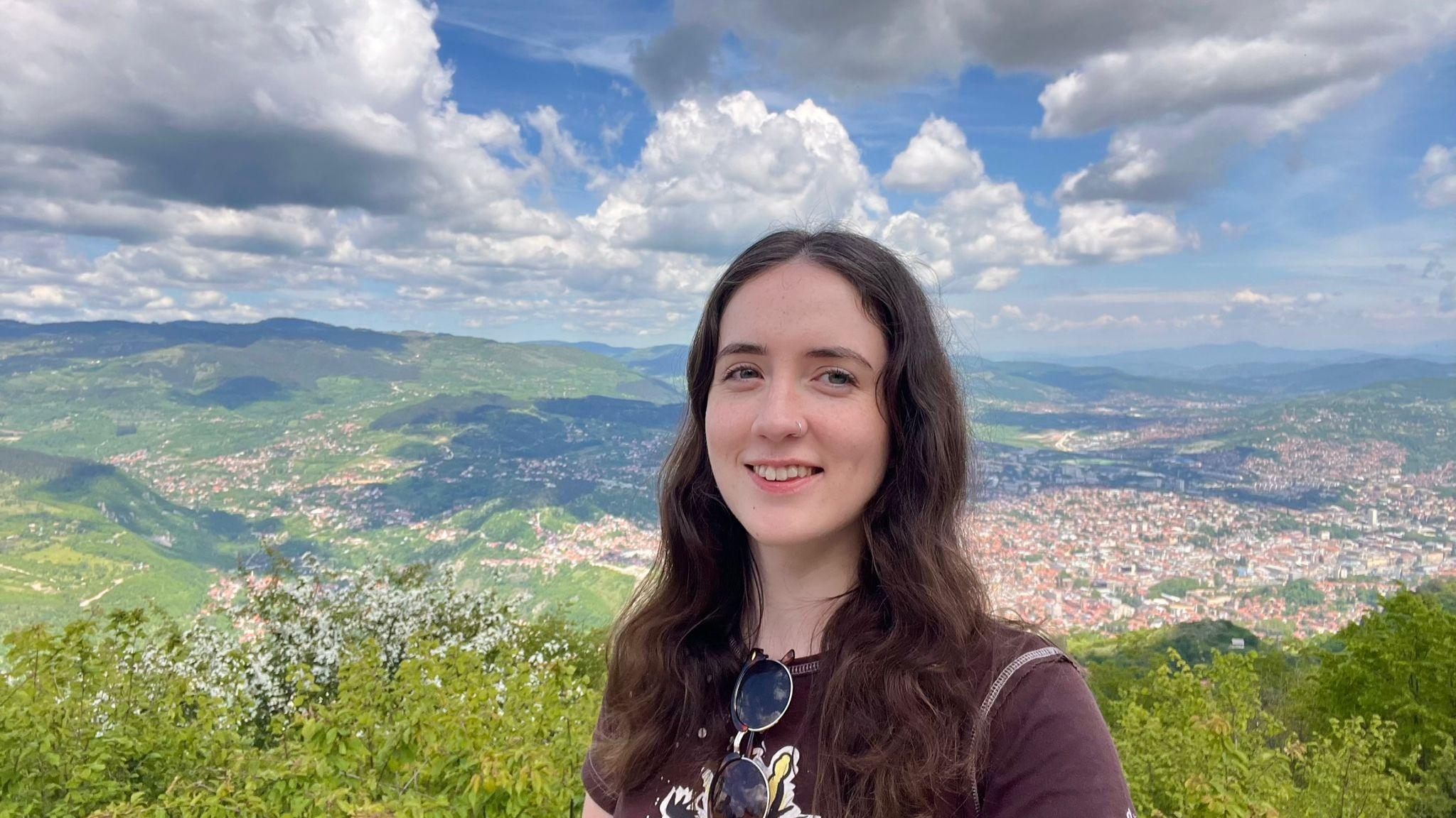 Em, një grua e re me flokë të errët. Ajo po qëndron në një rrugë të madhe përpara një peizazhi të madh të gjelbër me një qytet në distancë në të djathtë.