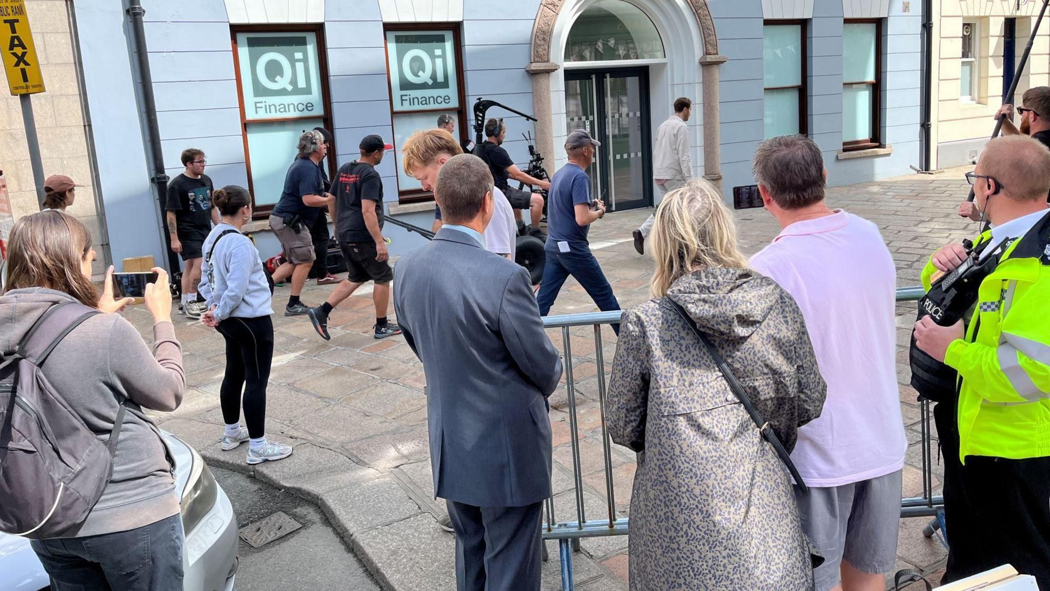 A film crew in the town of St Helier in Jersey watched by members of the public and police. An actor walks down a shopping street, followed by various members of the crew including a mounted camera operator.