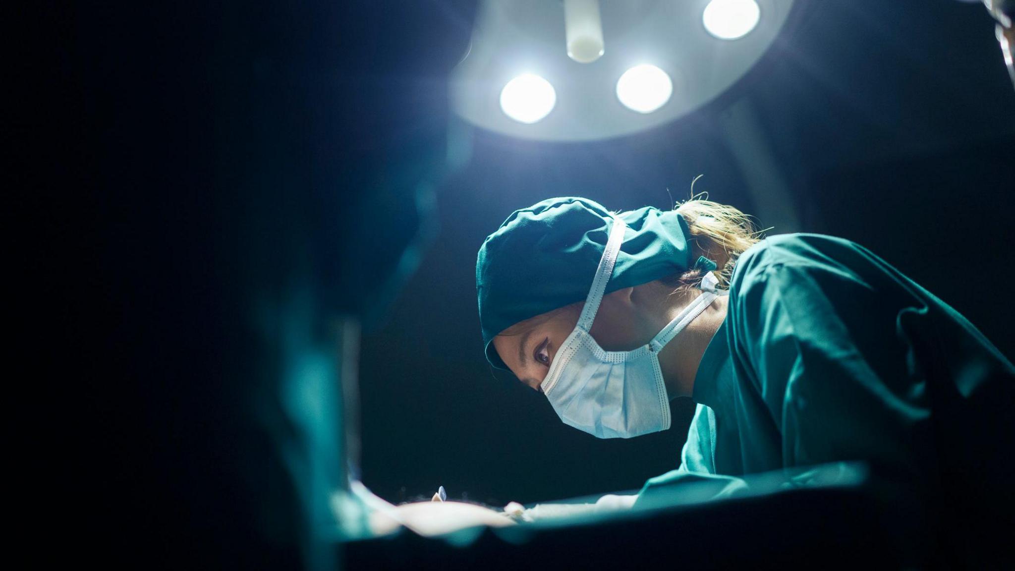 Surgeon wearing green scrubs and a face mask while working under a bright light.