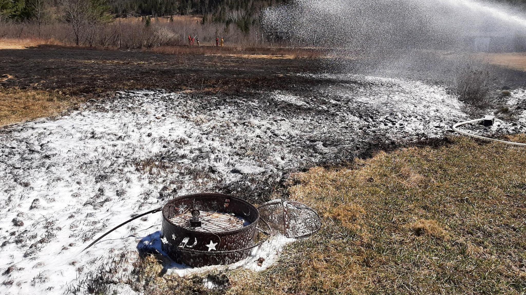 A grass field with a pine forest in the back appears charred. There is a burnt out barbeque and foam is being sprayed over the burnt area from the right of the picture. Part of the ground is covered in the firefighting foam.