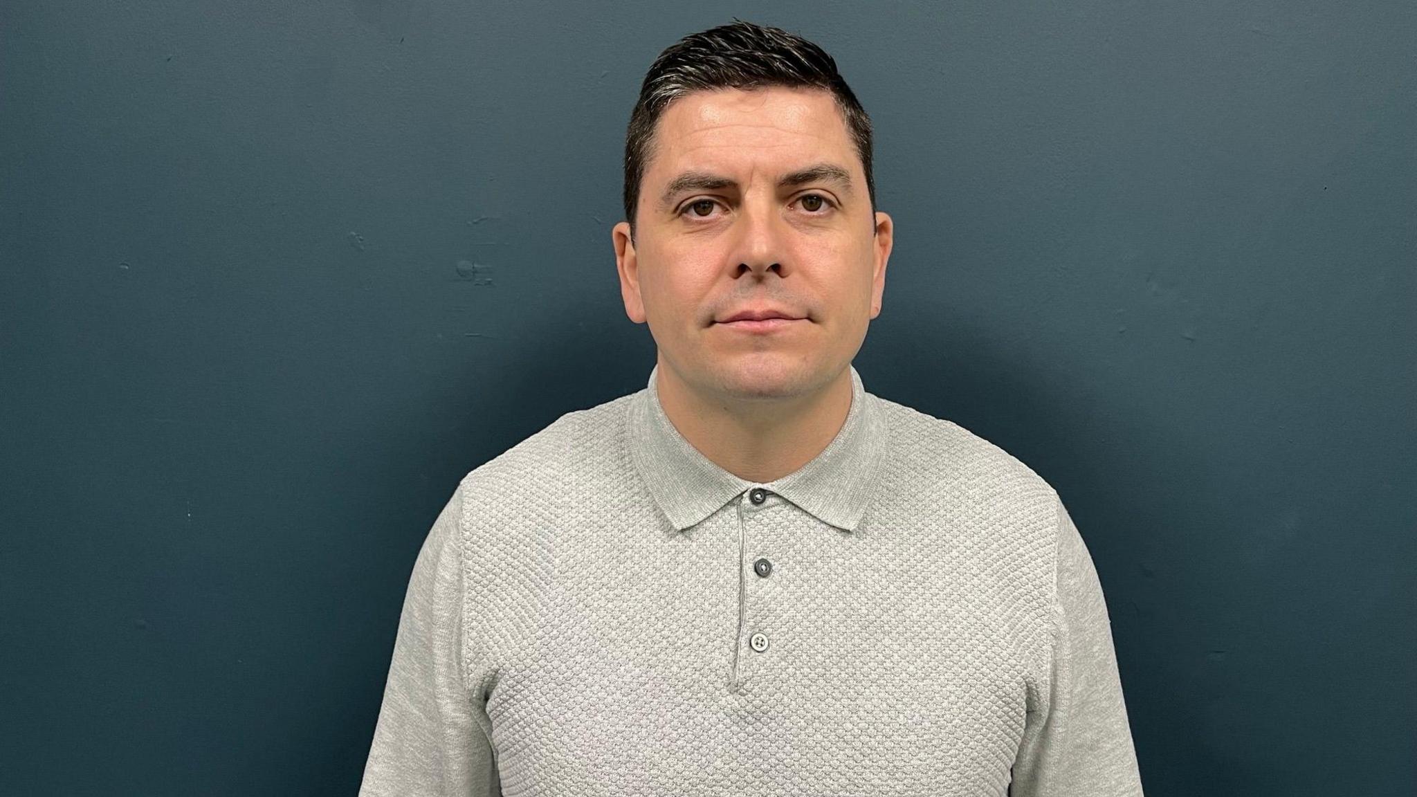 A man wearing a grey polo top, he has black hair and is standing in front of a dark green wall