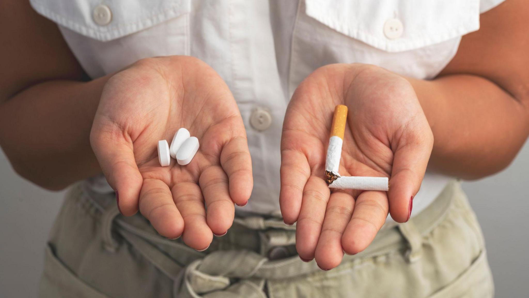 A young woman holds a few tablets in one palm and a broken, unlit cigarette in her other. 