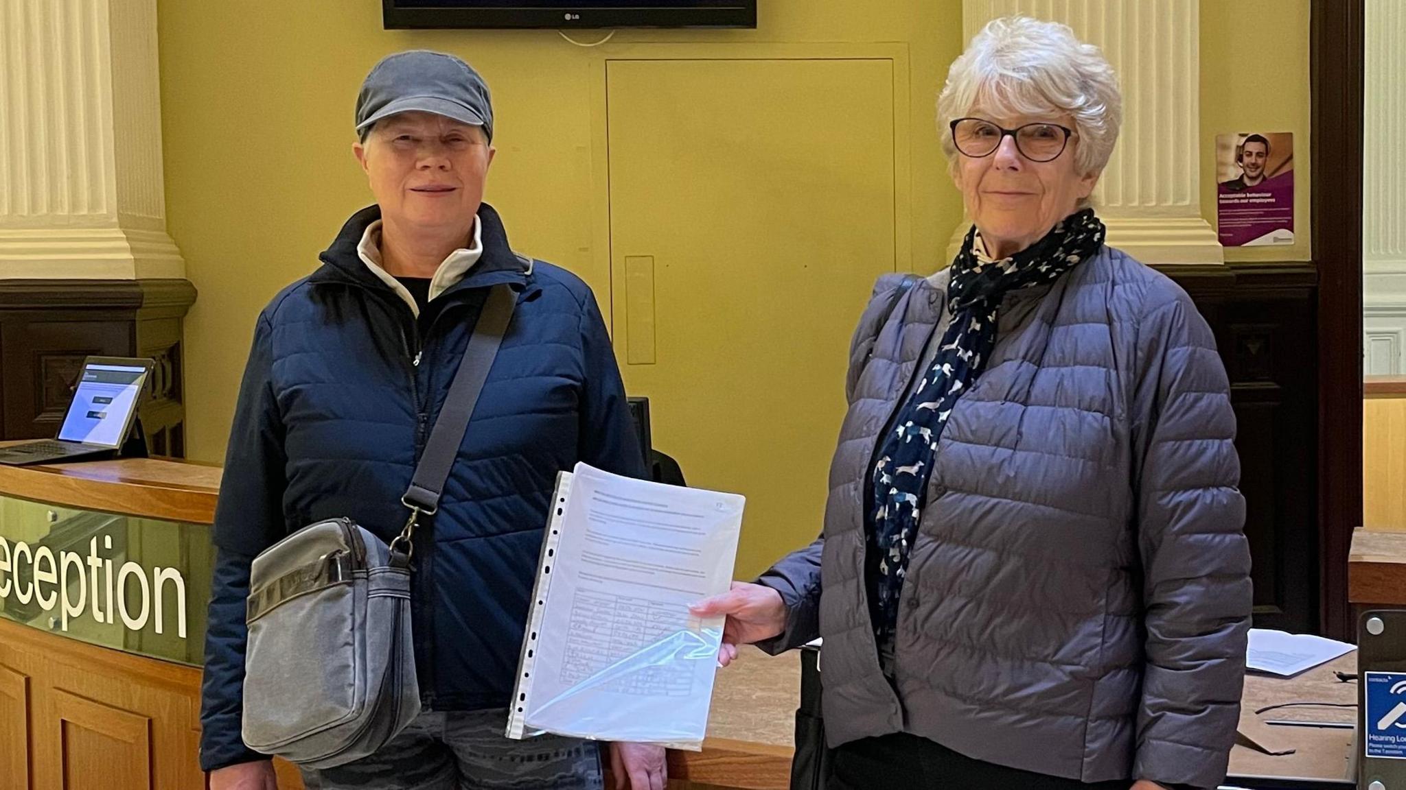 A photograph of two campaigners stood next to one another at the reception of the council's headquarters in Matlock. The lady on the right is holding the petition in her hand 