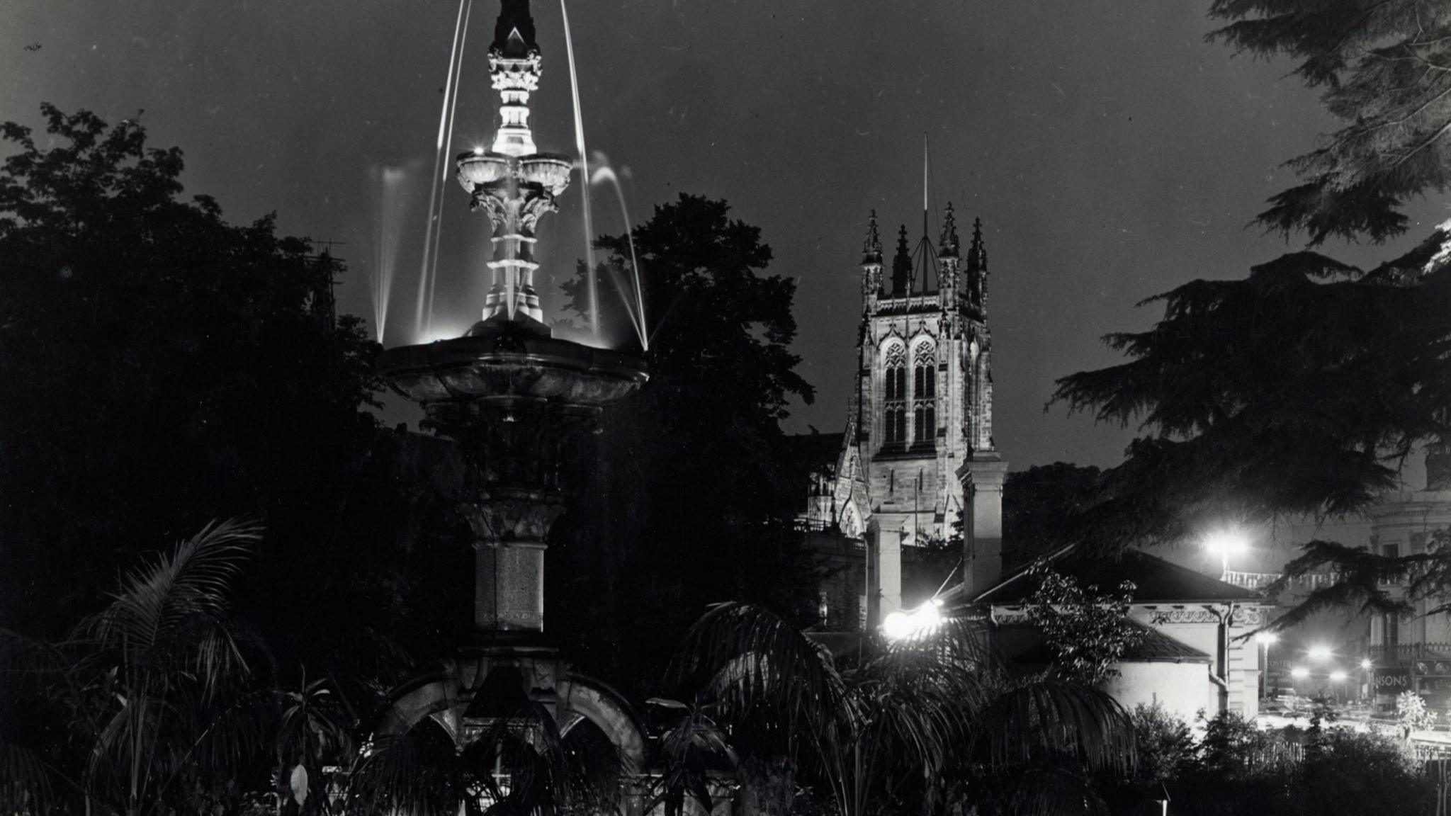 A black and white image showing an illuminated fountain surrounded by bushes and trees with a church in the background.