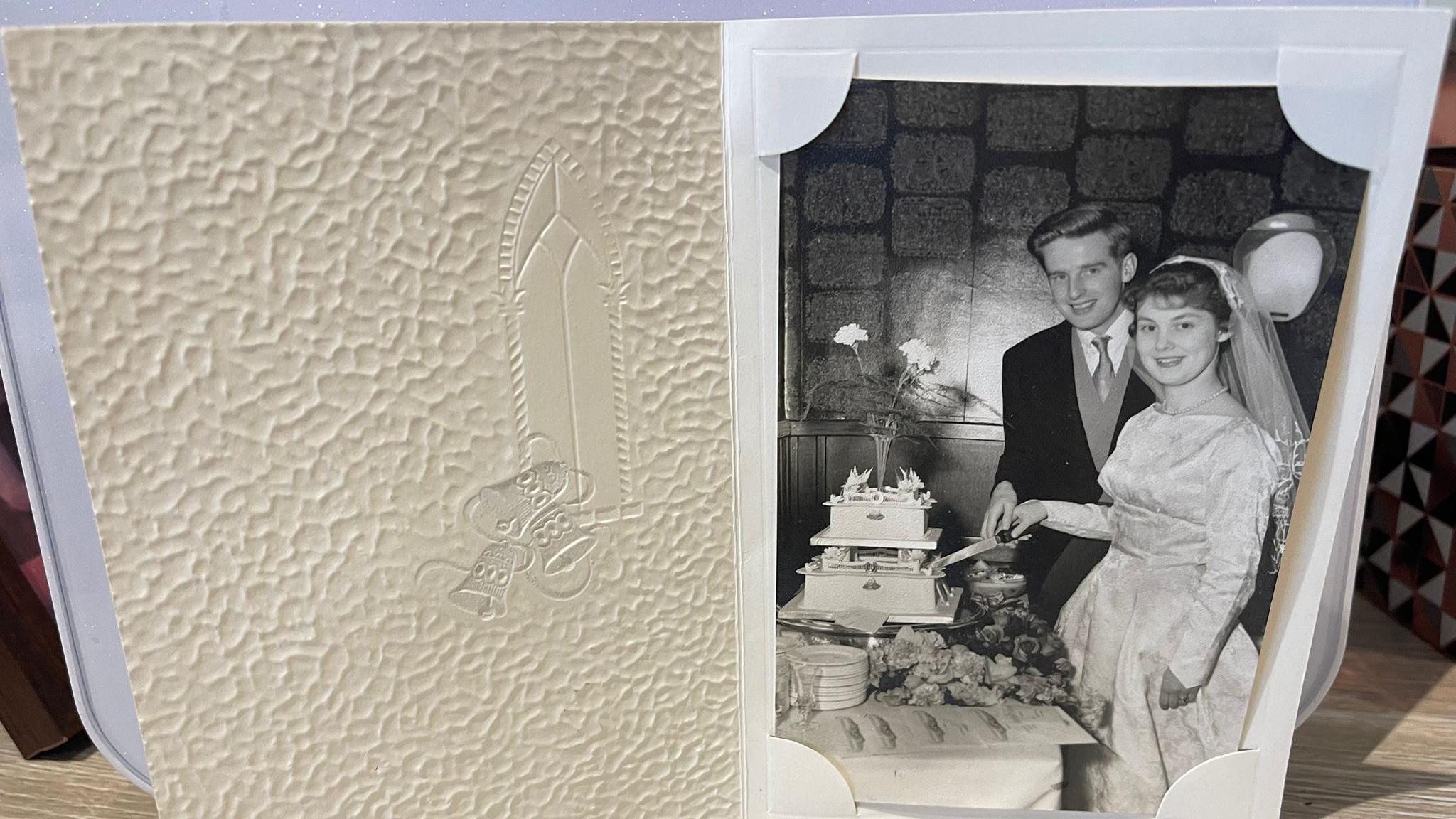 A card with a black and white photograph in showing Eileen and Peter on their wedding day cutting a two tiered cake.