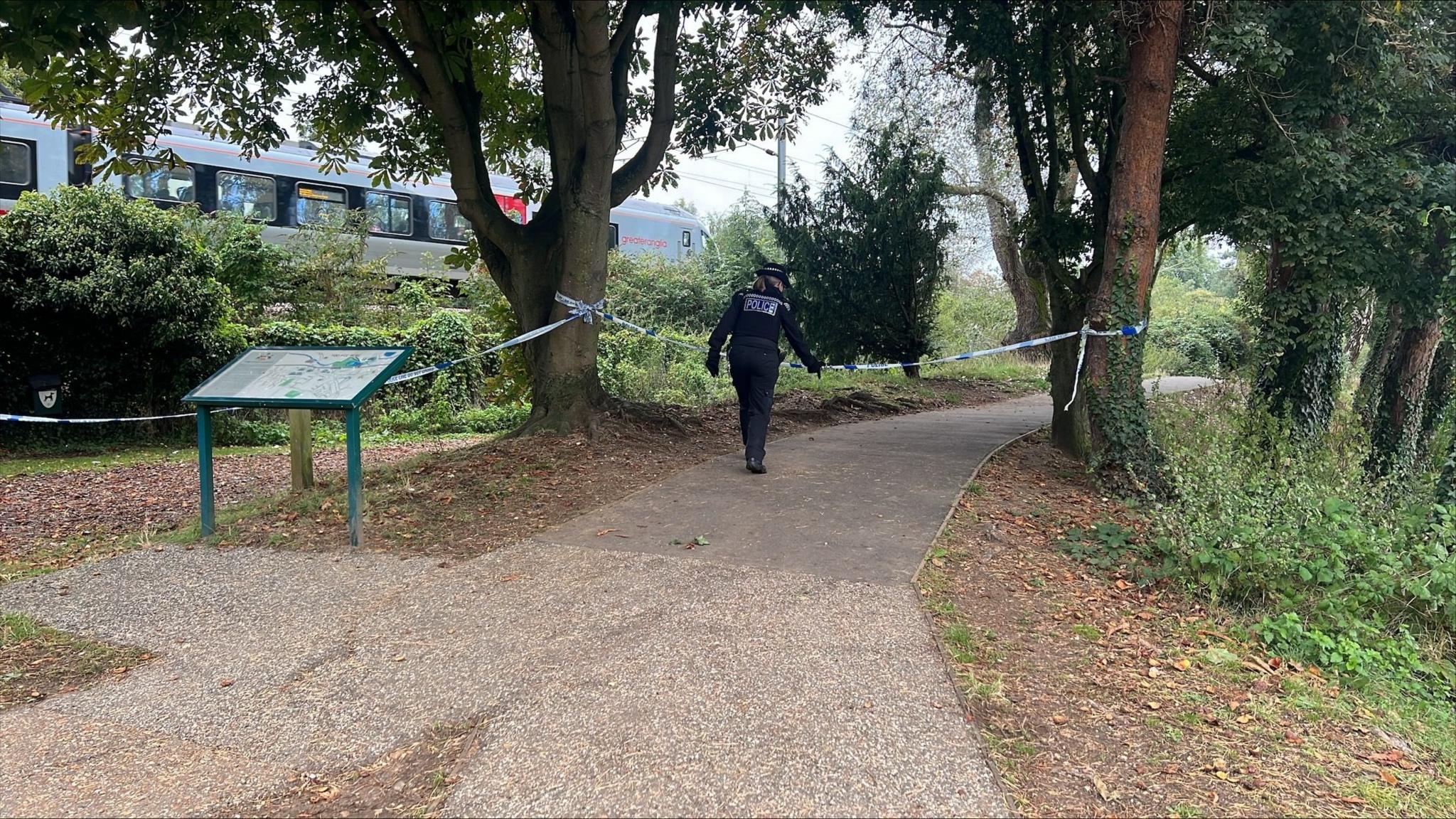 A general view of Needham Lake in Needham Market Suffolk. A footpath can be seen cordoned off with police tape while an officer can be seen in the background. An information board can be seen as well as trees.